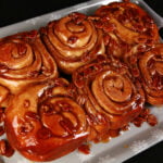 A close up view of a slab of 6 egg nog sticky buns. They are dripping with caramel and pecans.