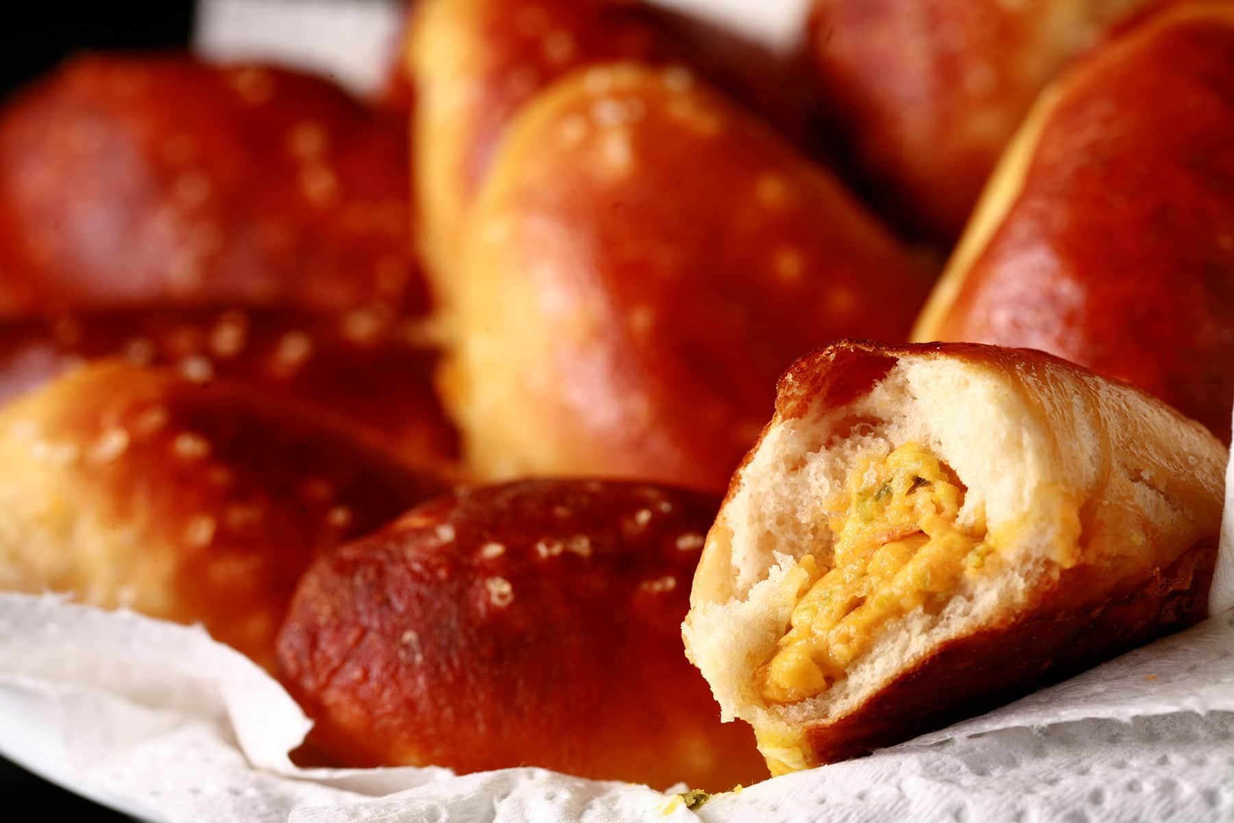 A napkin lined plastic food basket is full of golden brown soft pretzel bites. In the foreground, one of the buns has a bite taken out, revealing a jalapeno and cheese filling.