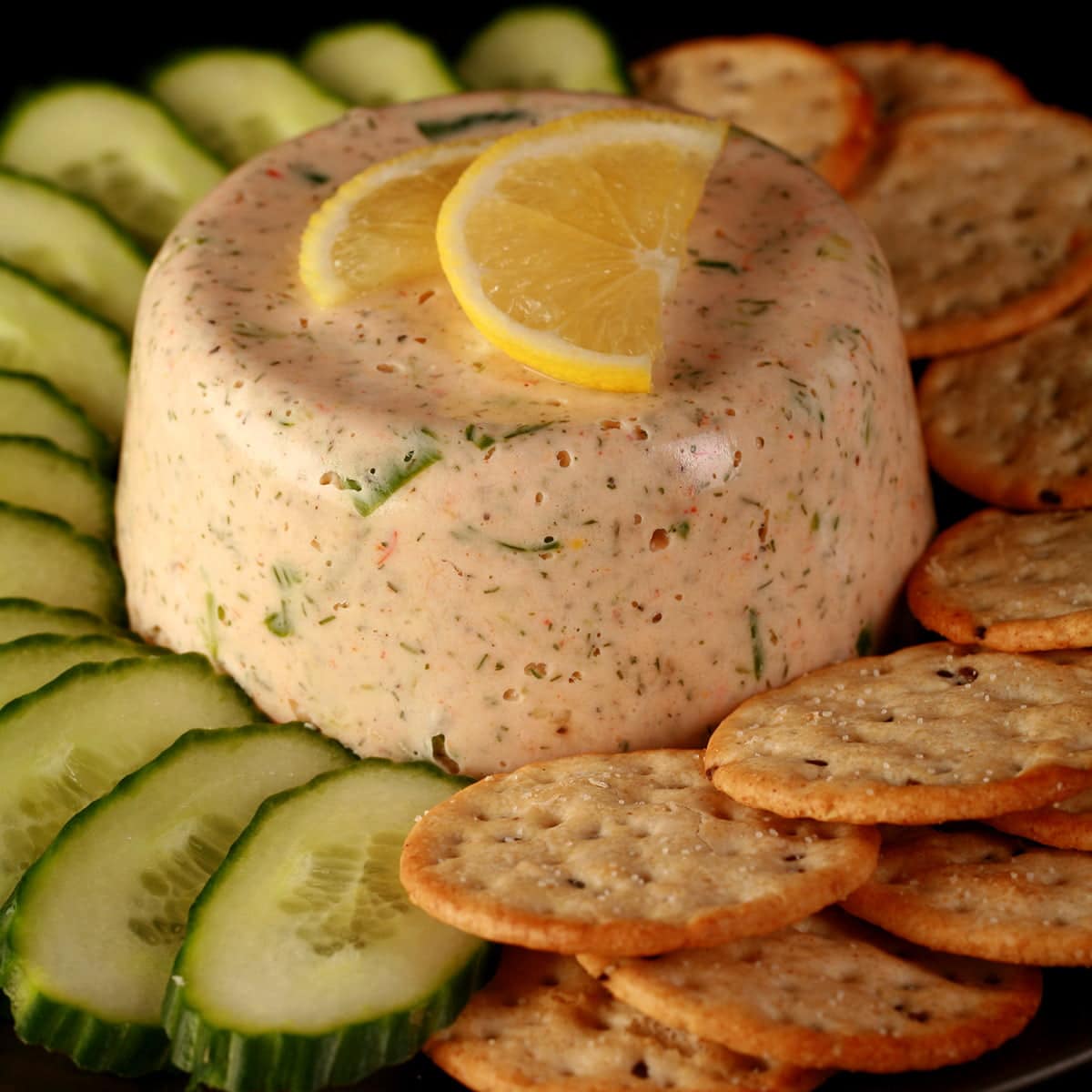 A closeup view of crab mousse. It is topped with slices of lemon and a sprig of parsley, and is surrounded by cucumber slices and crackers.