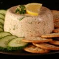 A closeup view of shrimp mousse. It is topped with slices of lemon and a sprig of parsley, and is surrounded by cucumber slices and crackers.