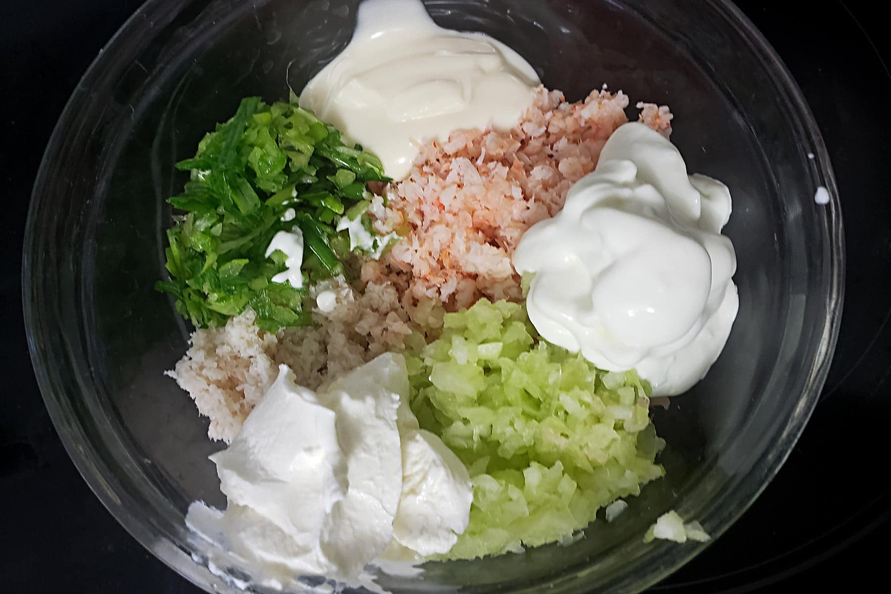 A glass bowl holds the ingredients of this seafood mousse recipe: canned seafood, celery, green onion, mayo, sour cream, and whipped cream.
