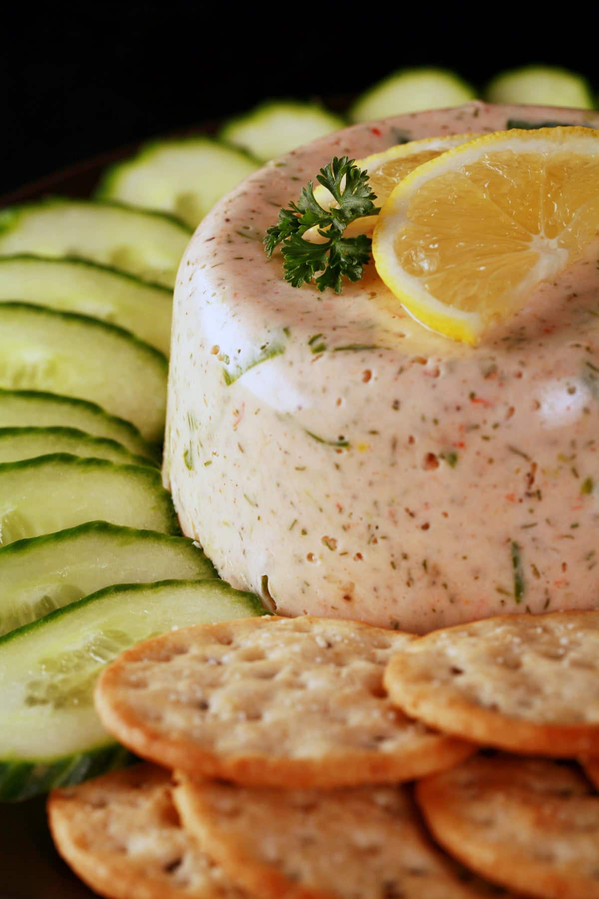 A closeup view of seafood mousse. It is topped with slices of lemon and a sprig of parsley, and is surrounded by cucumber slices and crackers.