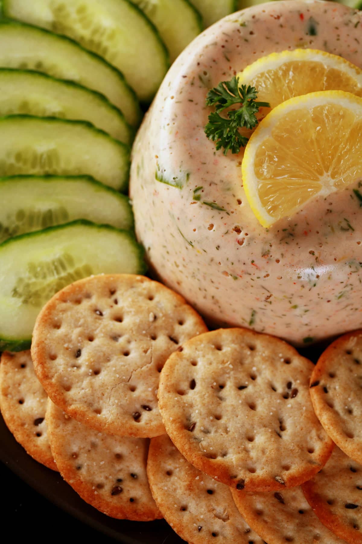 A closeup view of shrimp mousse. It is topped with slices of lemon and a sprig of parsley, and is surrounded by cucumber slices and crackers.