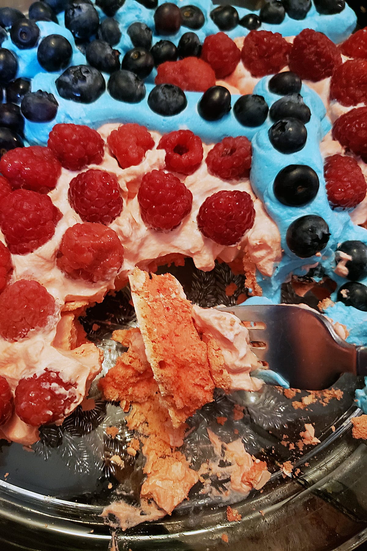 A close up view of a partially eaten red and blue pavlova, with raspberries and blueberries.