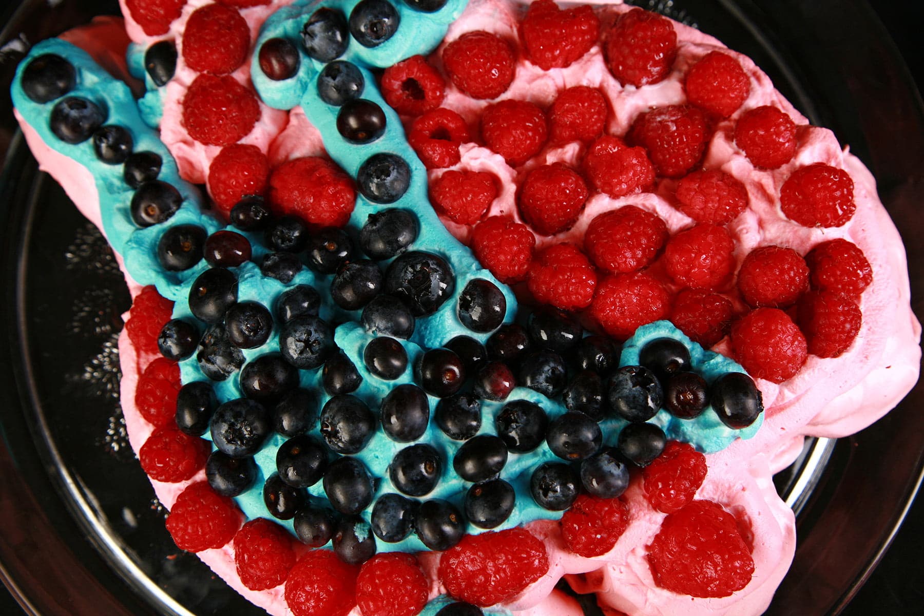 A close up view of a pink pavlova, with pink whipped cream, blue whipped cream, blueberries, and raspberries.