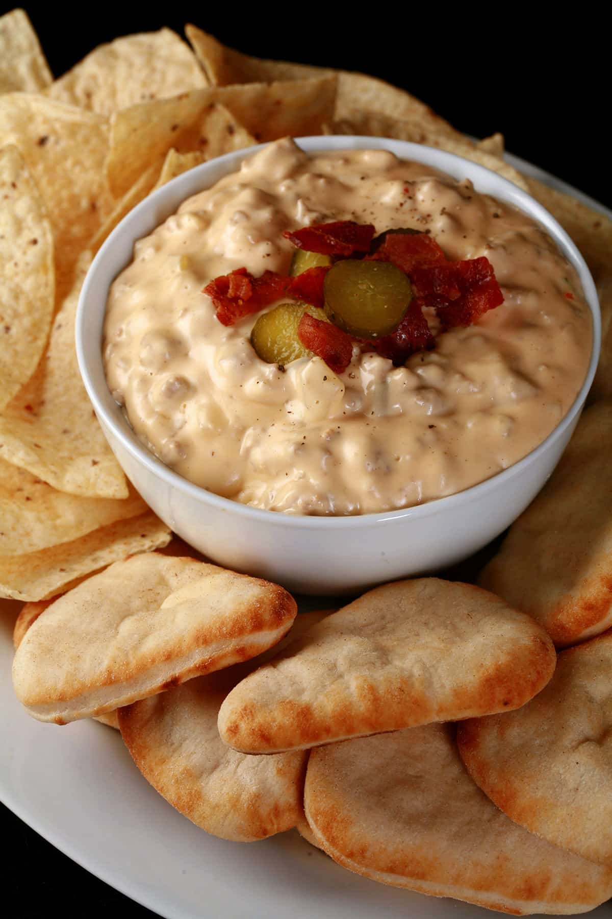 A bowl of hot cheeseburger dip, topped with bacon and pickles, and surrounded by pita bites and corn tortilla chips.