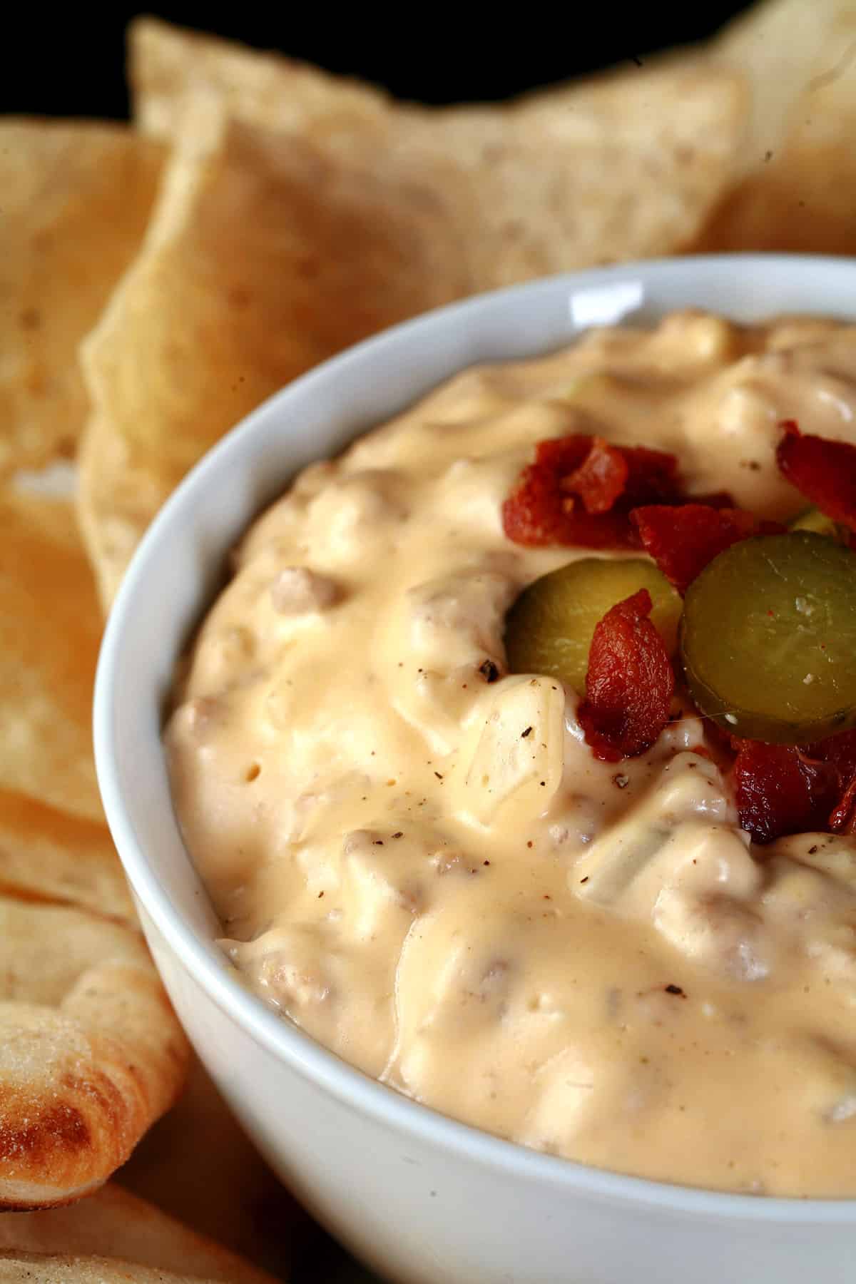 A bowl of hot cheeseburger dip, topped with bacon and pickles, and surrounded by pita bites and corn tortilla chips.