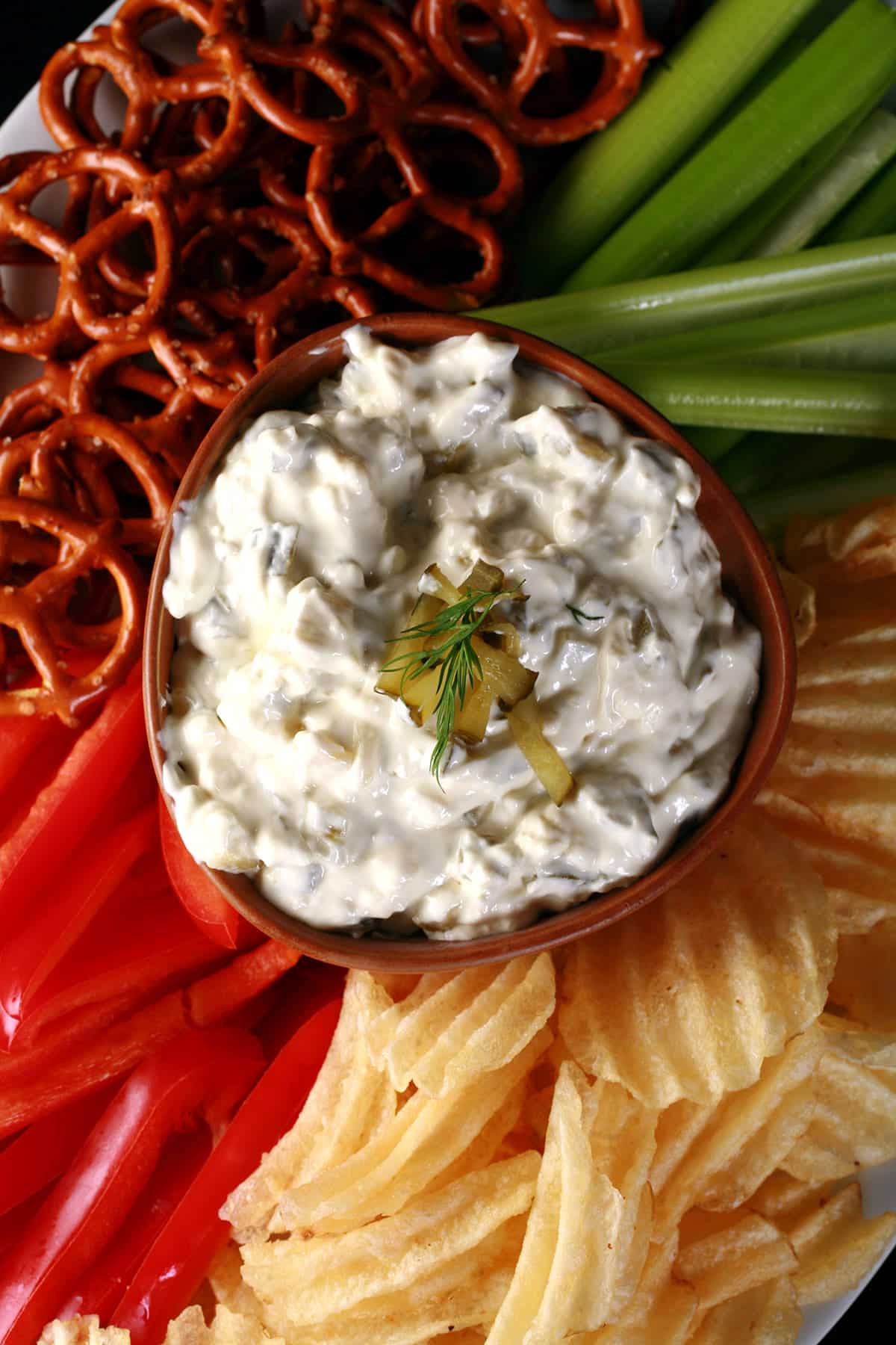 A bowl of dill pickle dip, surrounded with chips, pretzels, and cut veggies.