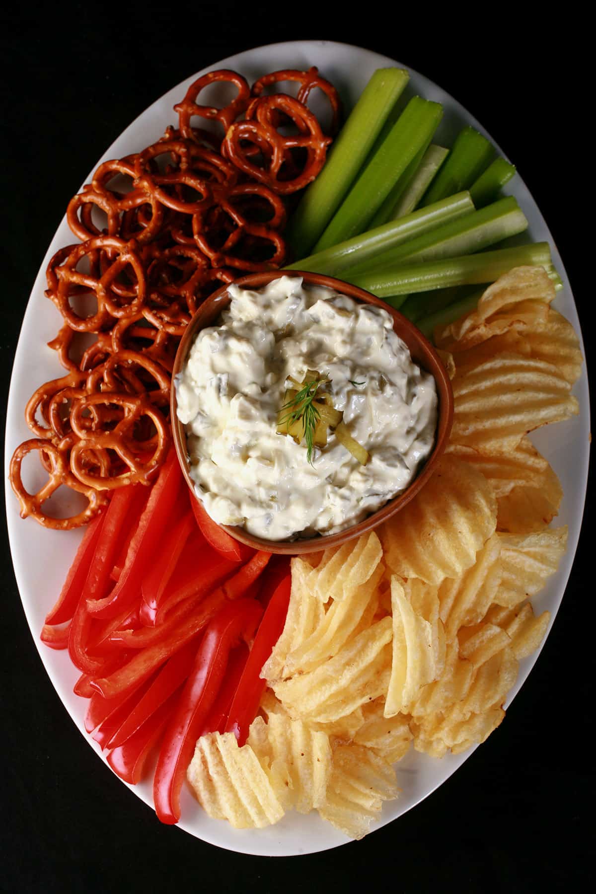 A bowl of dill pickle cream cheese dip - based on the Phiadelphia Cream Cheese brand dip - surrounded with chips, pretzels, and vegetables.