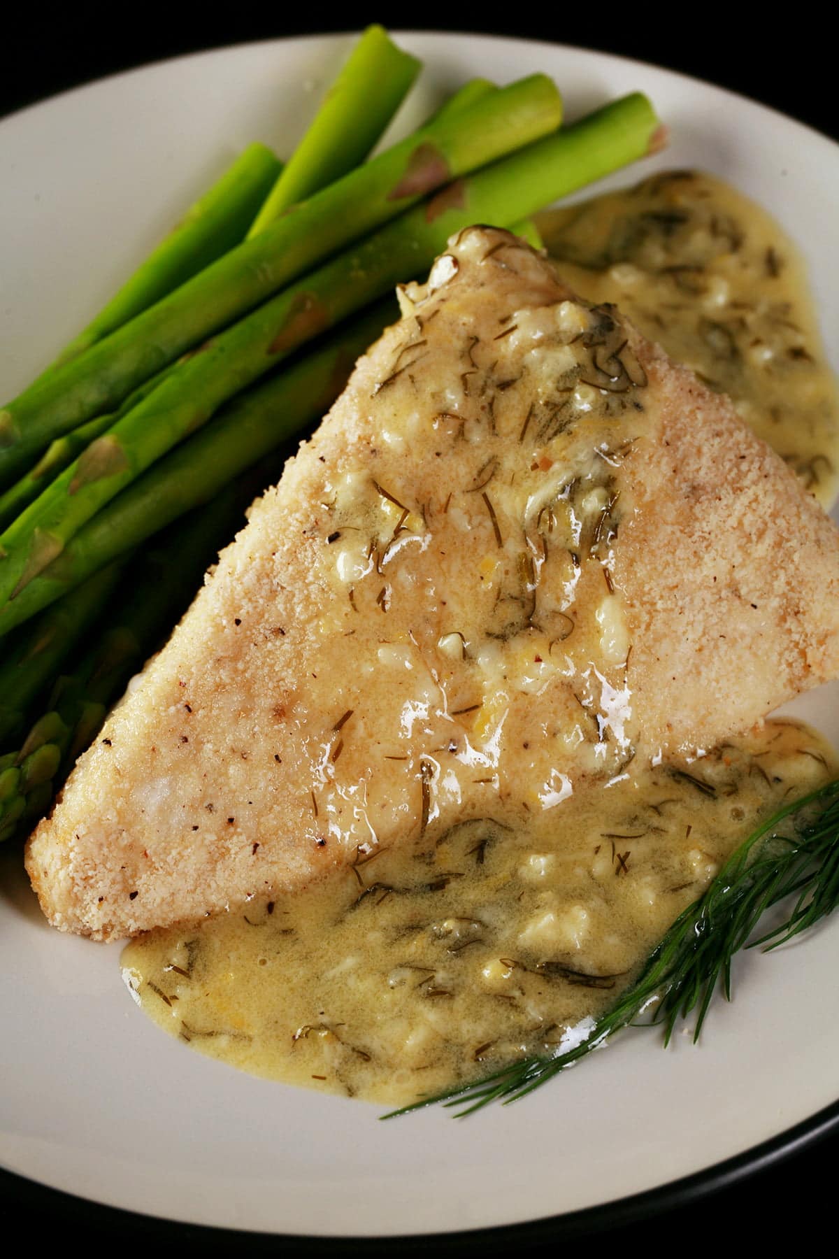 A large piece of almond crusted halibut is on a white plate. A yellow cream sauce with flecks of green - lemon dill cream sauce - has been poured over the fish, and there are asparagus spears on the side.
