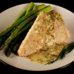 A large piece of almond crusted halibut is on a white plate. A yellow cream sauce with flecks of green - lemon dill cream sauce - has been poured over the fish, and there are asparagus spears on the side.