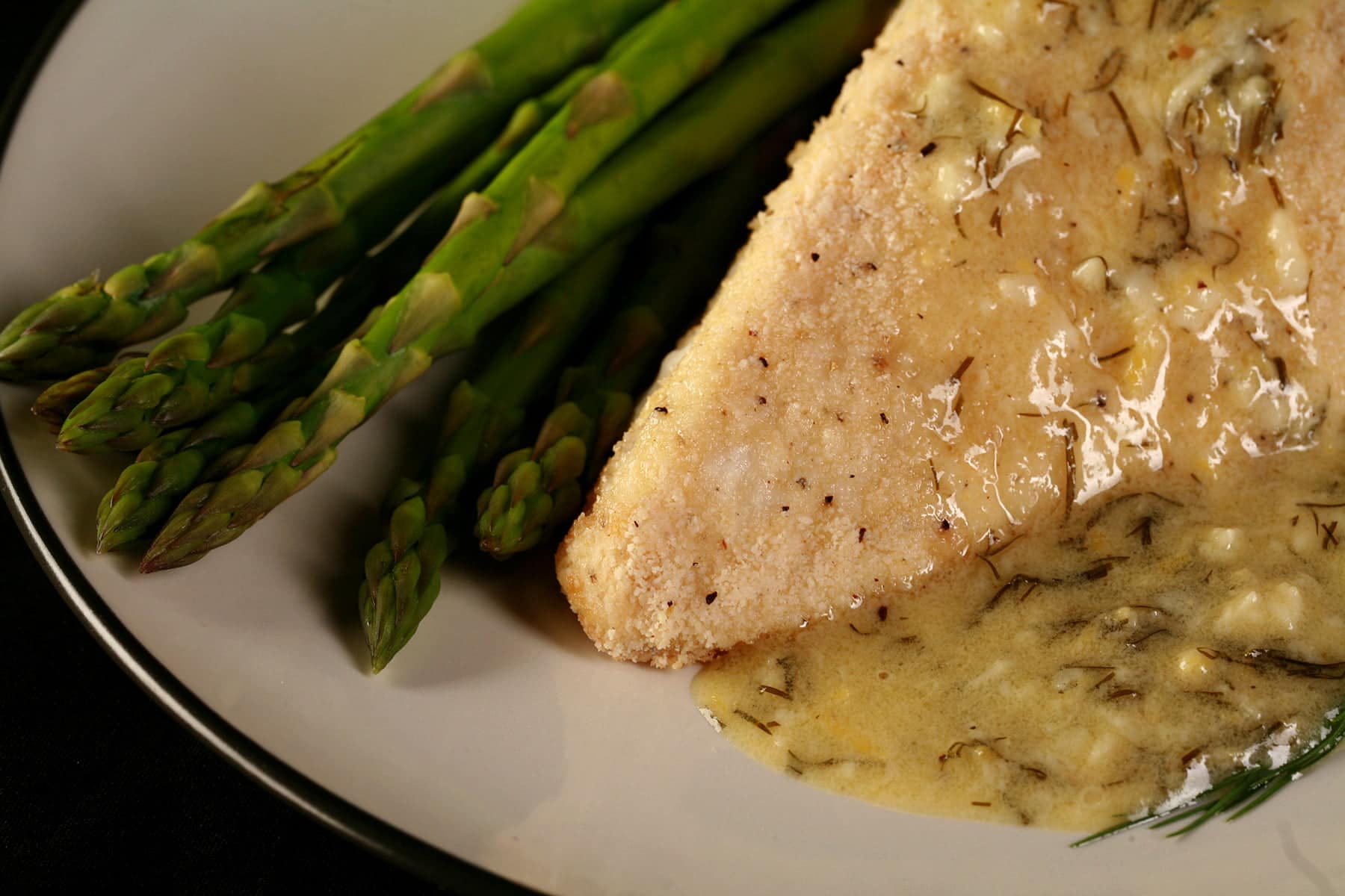 A large piece of almond crusted halibut is on a white plate. A yellow cream sauce with flecks of green - lemon dill cream sauce - has been poured over the fish, and there are asparagus spears on the side.