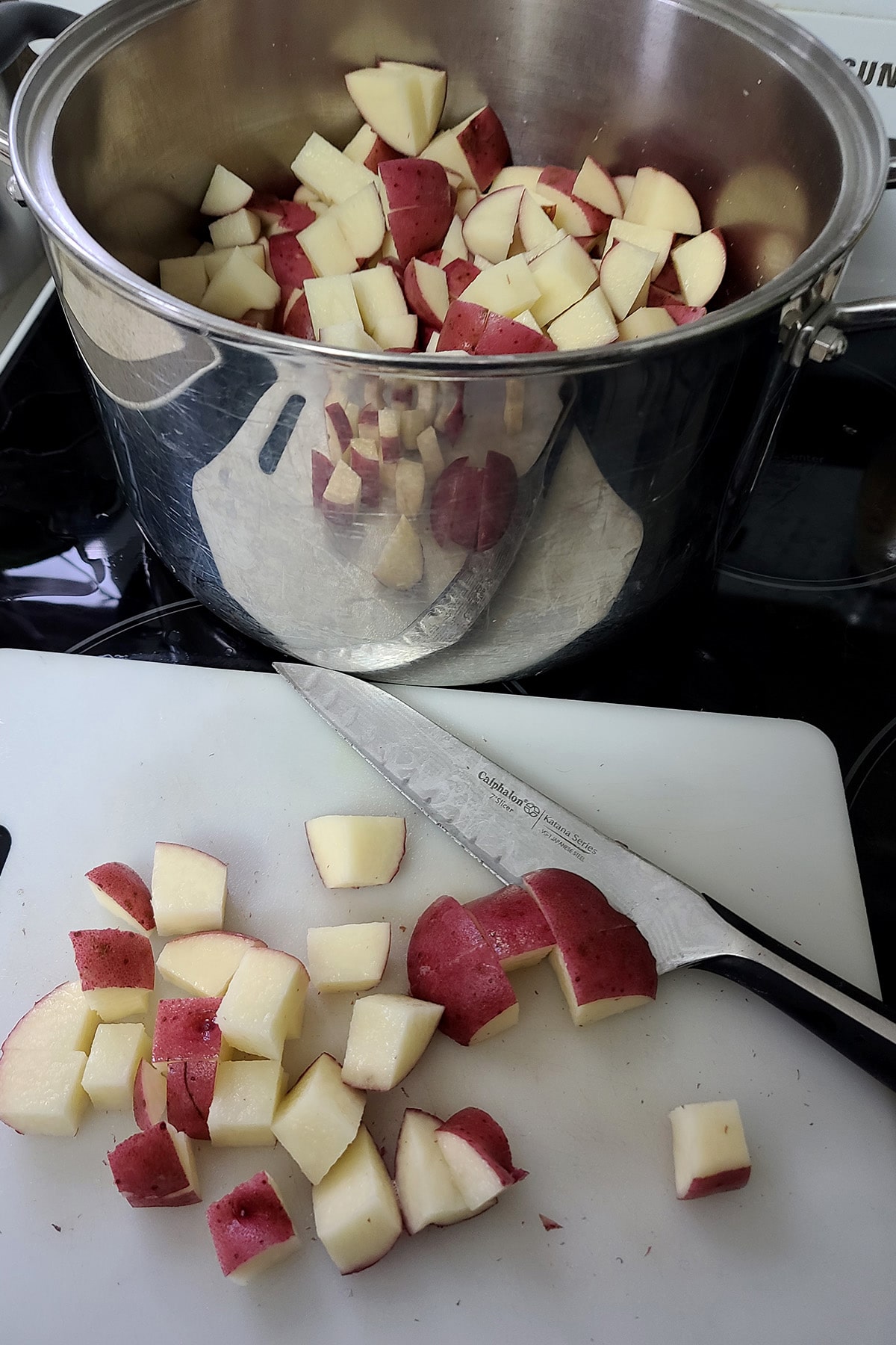 A cutting board with chopped red potatoes on it, in front of a large pot with chopped potatoes in it.