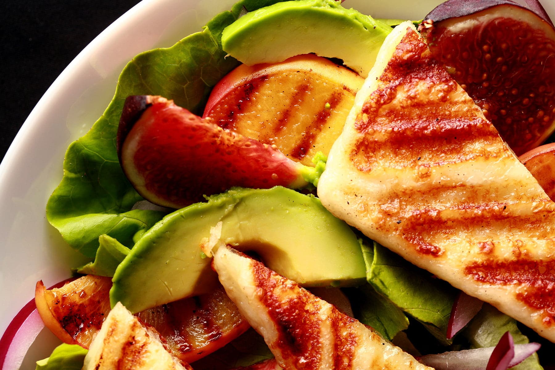 A bowl of salad, with sliced figs, grilled peaches, and strips of grilled white cheese. Grilled Halloumi Salad with Peaches and Figs