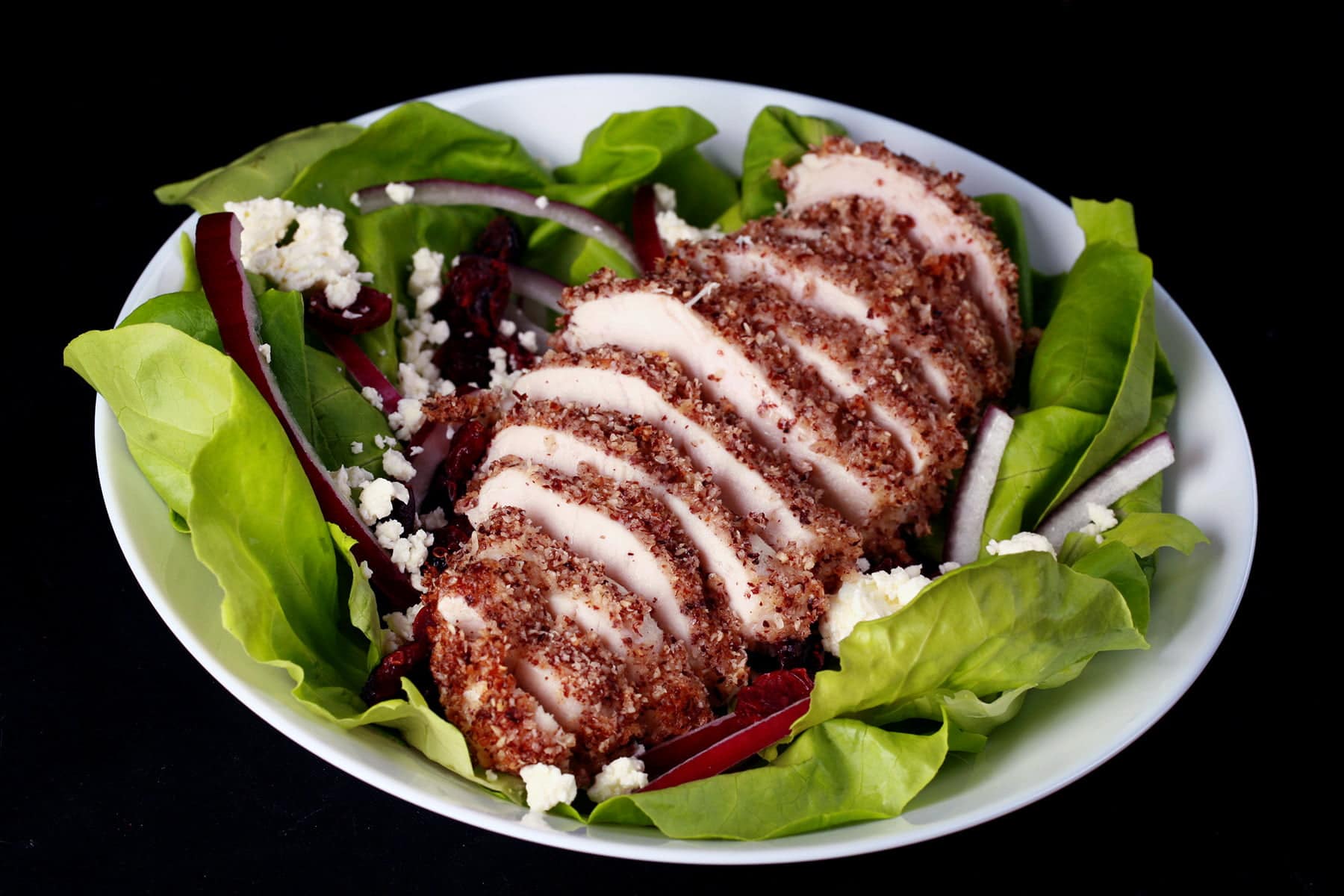 A sliced hazelnut crusted chicken breast sits a top a salad of greens, pear slices, dried cranberries, red onion slices, and feta.