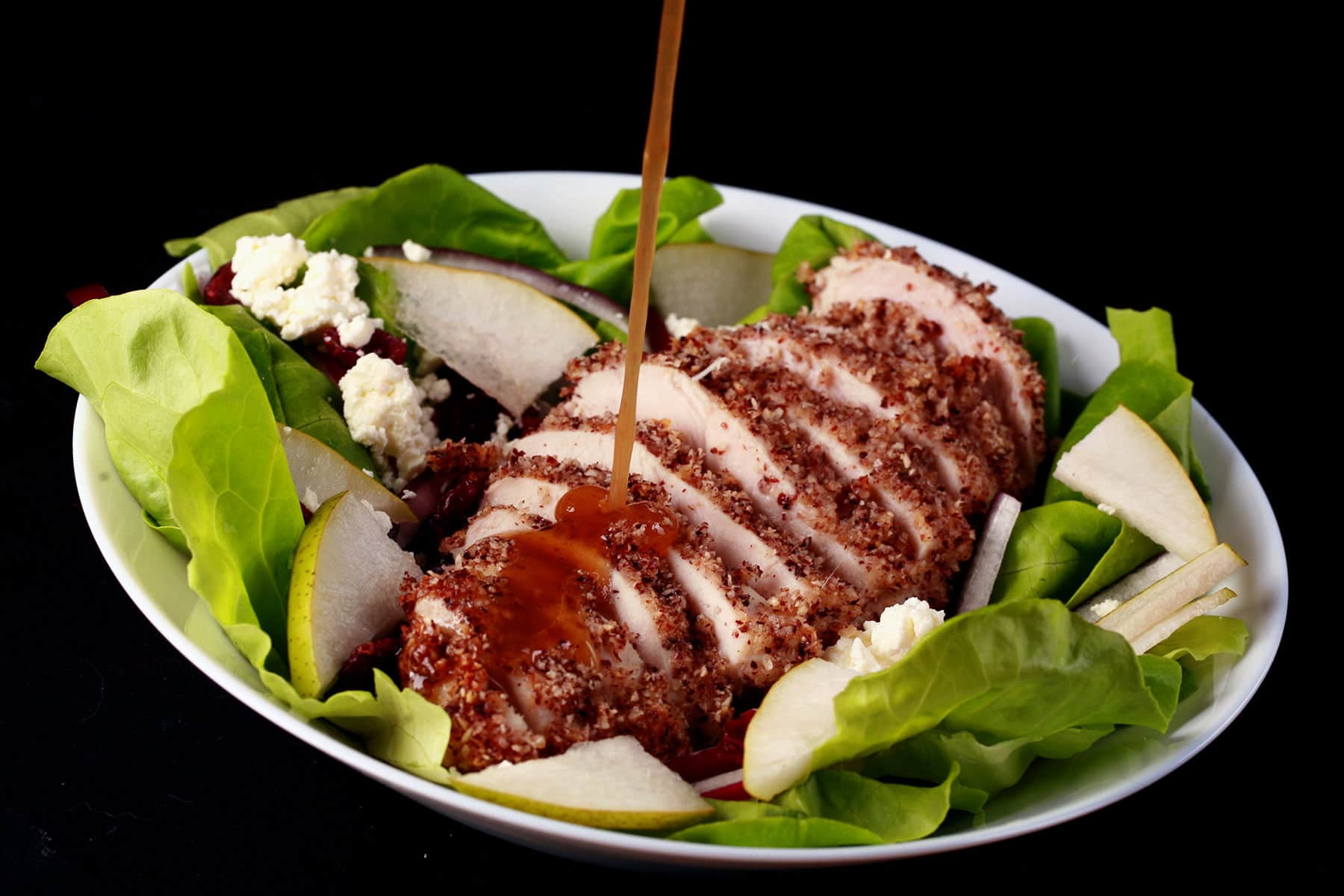 A sliced hazelnut crusted chicken breast sits a top a salad of greens, pear slices, dried cranberries, red onion slices, and feta. A golden brown balsamic vinaigrette is being poured across the chicken.