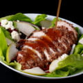 A sliced hazelnut crusted chicken breast sits a top a salad of greens, pear slices, dried cranberries, red onion slices, and feta. A golden brown balsamic vinaigrette is being poured across the chicken.