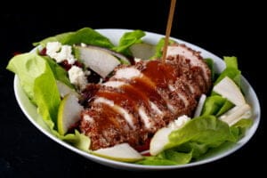 A sliced hazelnut crusted chicken breast sits a top a salad of greens, pear slices, dried cranberries, red onion slices, and feta. A golden brown balsamic vinaigrette is being poured across the chicken.
