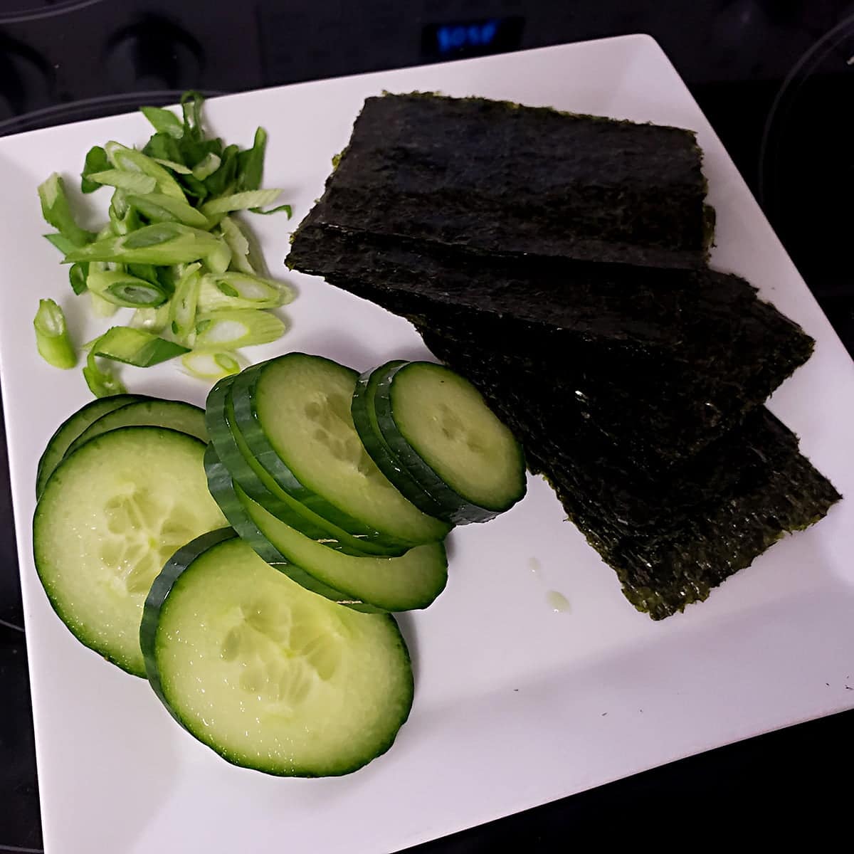 A white cutting board with slices of cucumber and green onions on it, along with pieces of nori - seaweed sheets.