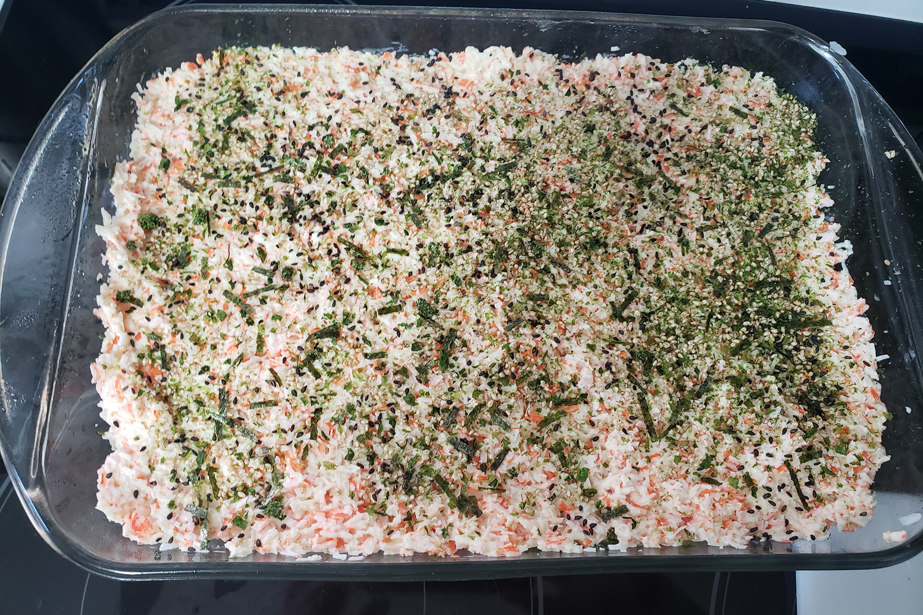 A large glass baking dish is shown with green furikake mix spread over the spicy crab mixture.