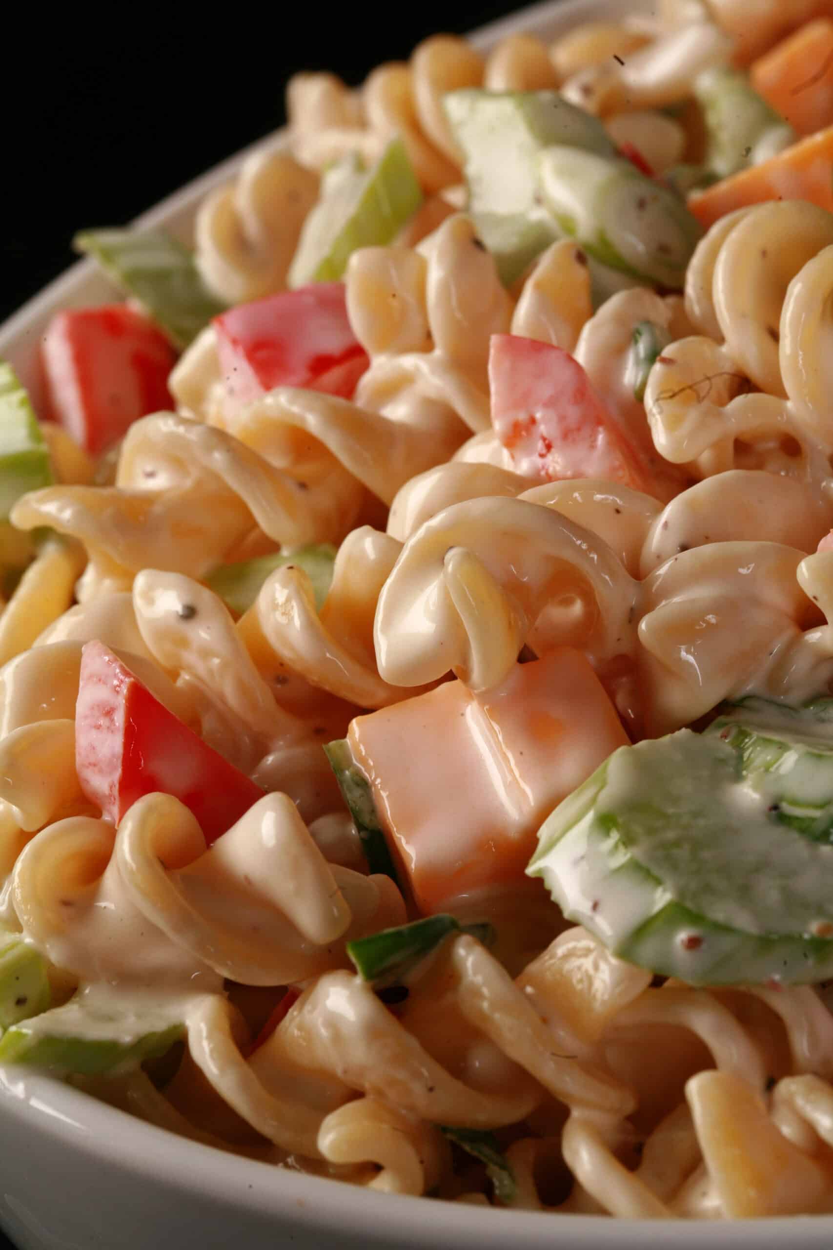 A close up view of a creamy pasta salad made with rotini, red peppers, celery, cheese, and green onions.