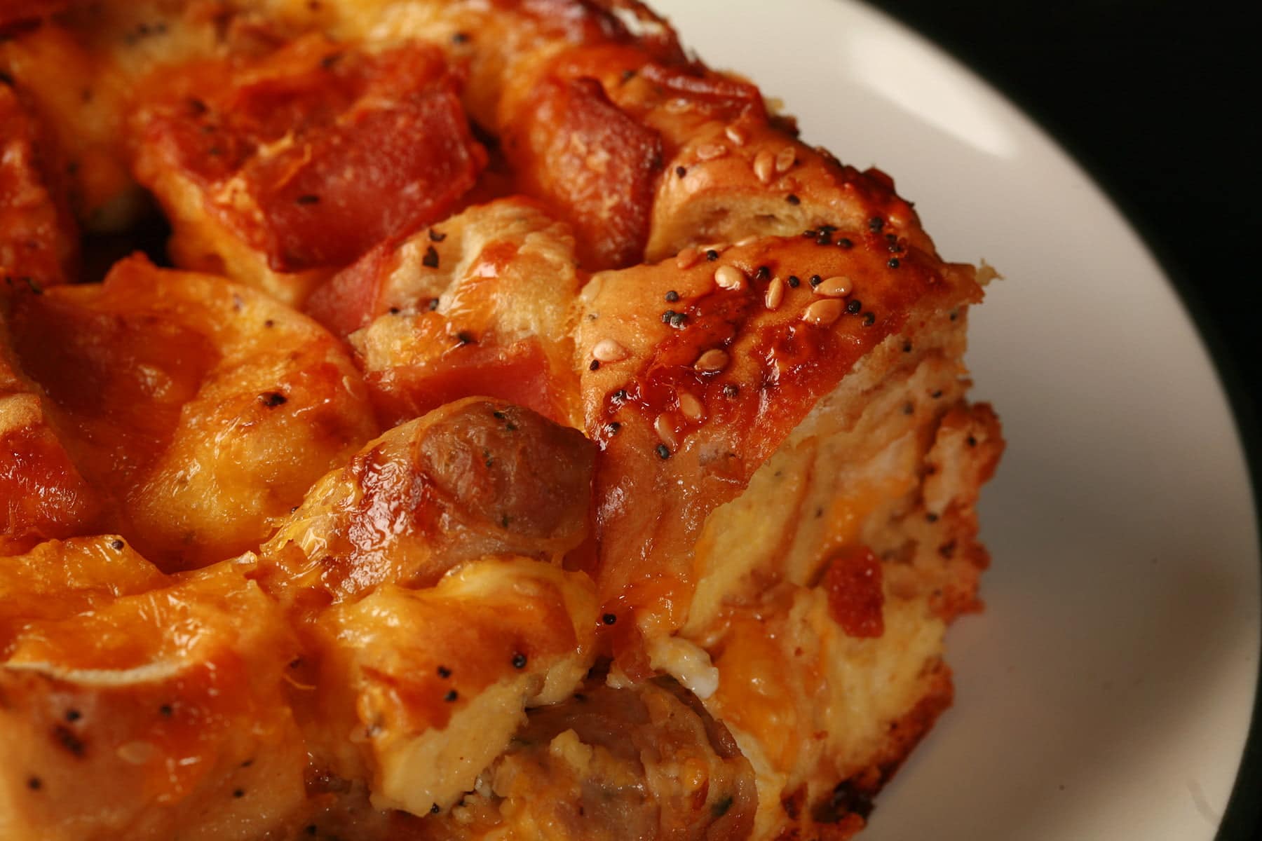 A square serving of breakfast bagel strata. Eggs, bacon, chunks of bagel, and pieces of sausage are visible in the cheesy casserole.