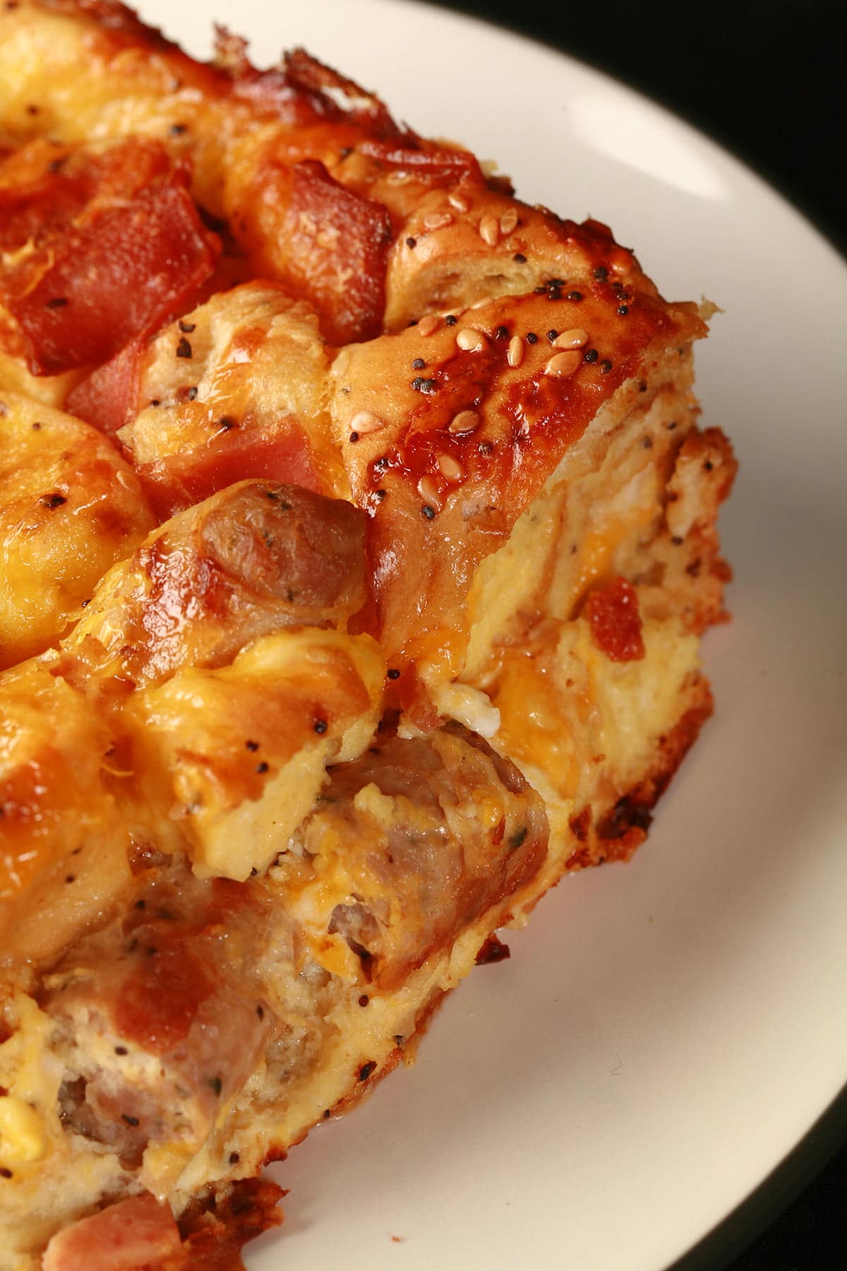 A square serving of breakfast bagel casserole. Eggs, bacon, chunks of bagel, and pieces of sausage are visible in the cheesy casserole.