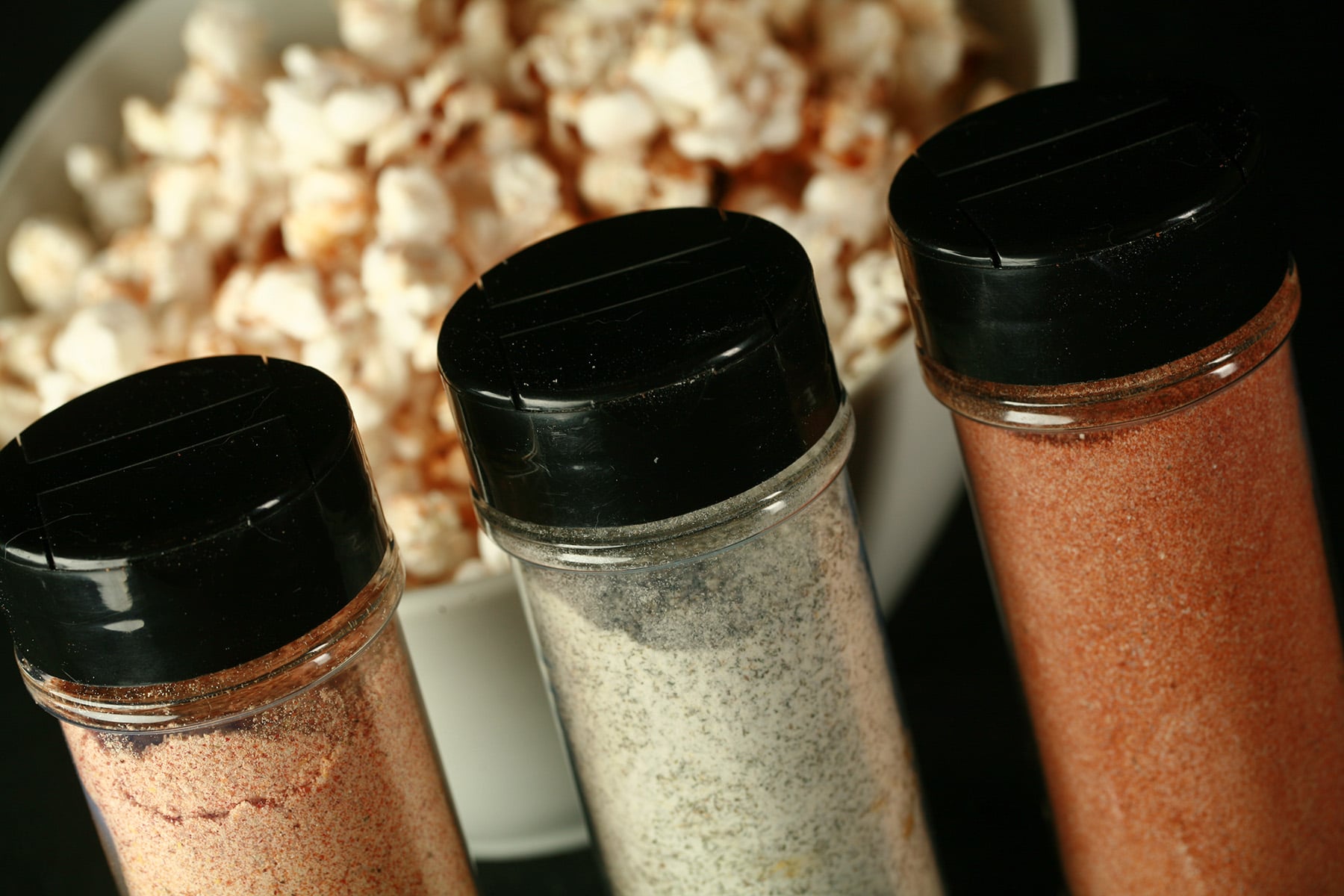 3 small canisters of popcorn seasoning - All Dressed, Dill Pickle, and Ketchup - are lined up in front of a large white bowl of seasoned popcorn.