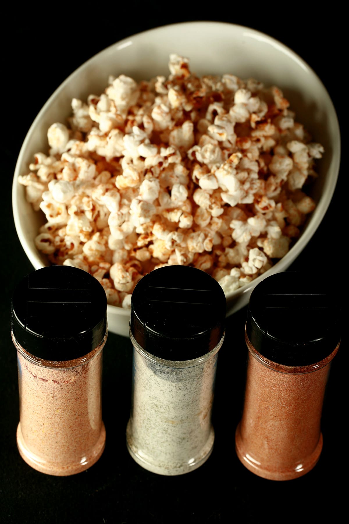 3 small canisters of popcorn seasoning - All Dressed, Dill Pickle, and Ketchup - are lined up in front of a large white bowl of seasoned popcorn.