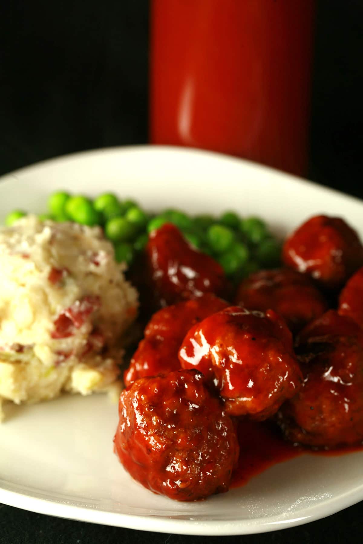 A plate of BBQ meatballs, mashed potatoes, and peas. The meatballs are coated in replica Original Diana Sauce. A bottle of the sauce rests behind the plate of food.