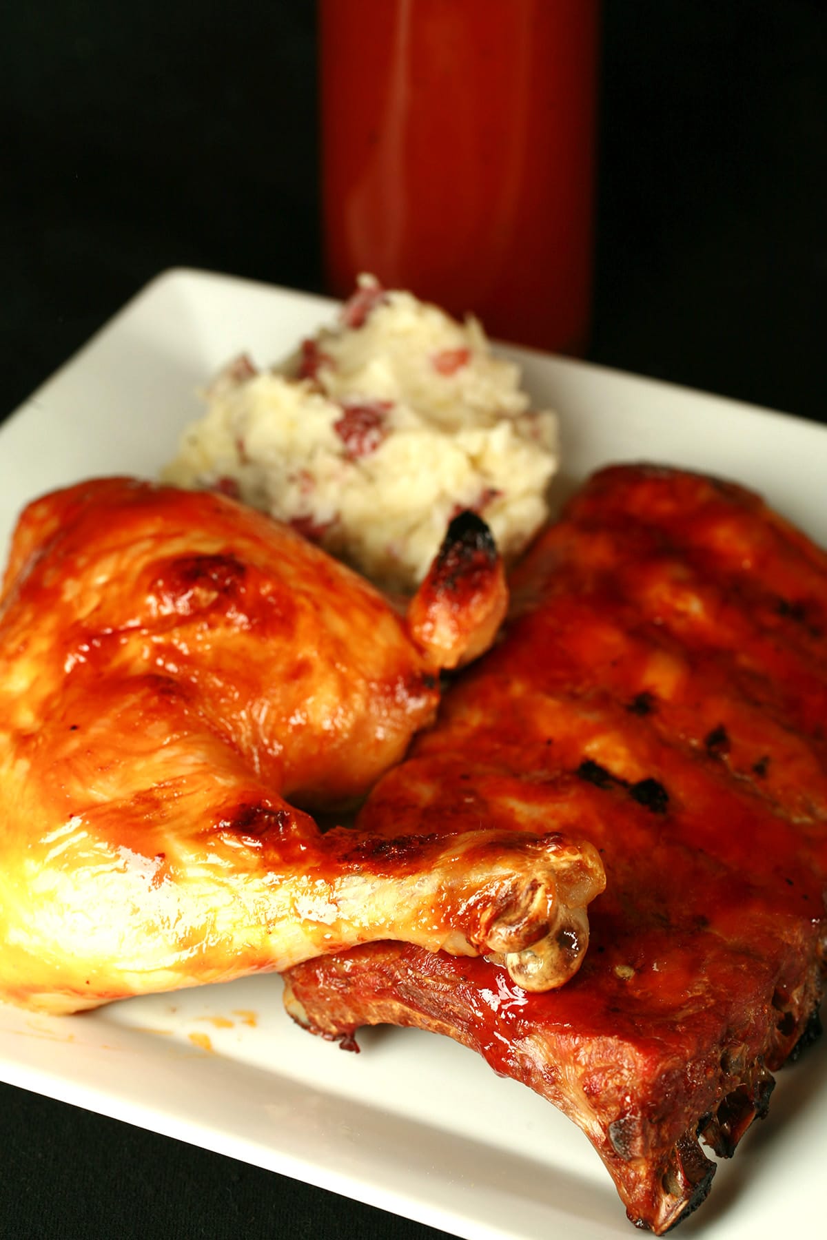 A plate of BBQ chicken, ribs, and mashed potatoes. The meats are coated in replica Diana Rib & Chicken Sauce. A bottle of the sauce rests behind the plate of food.