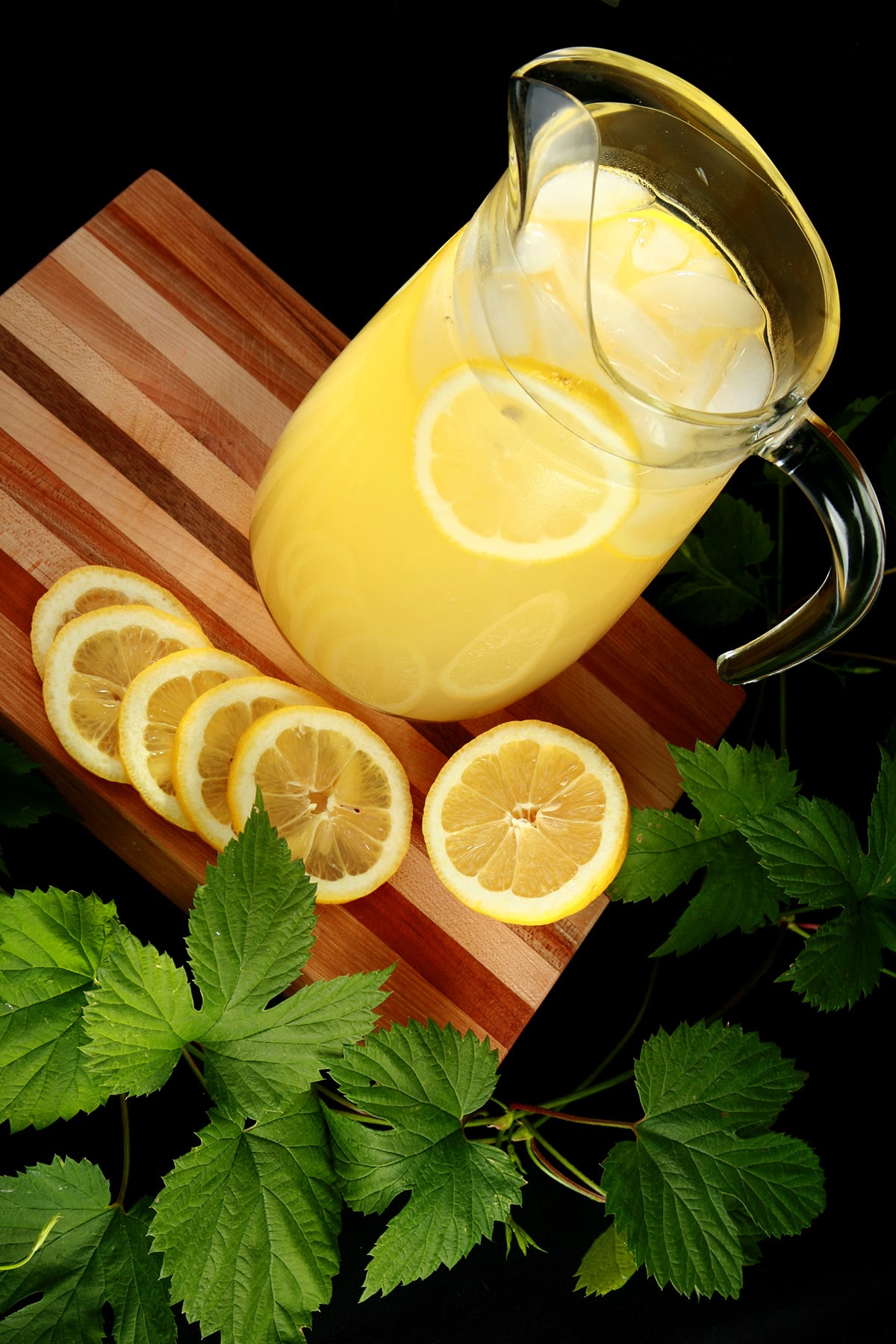 A pitcher of hop lemonade rests on a wooden board, with slices lemons and a hop bine next to it.