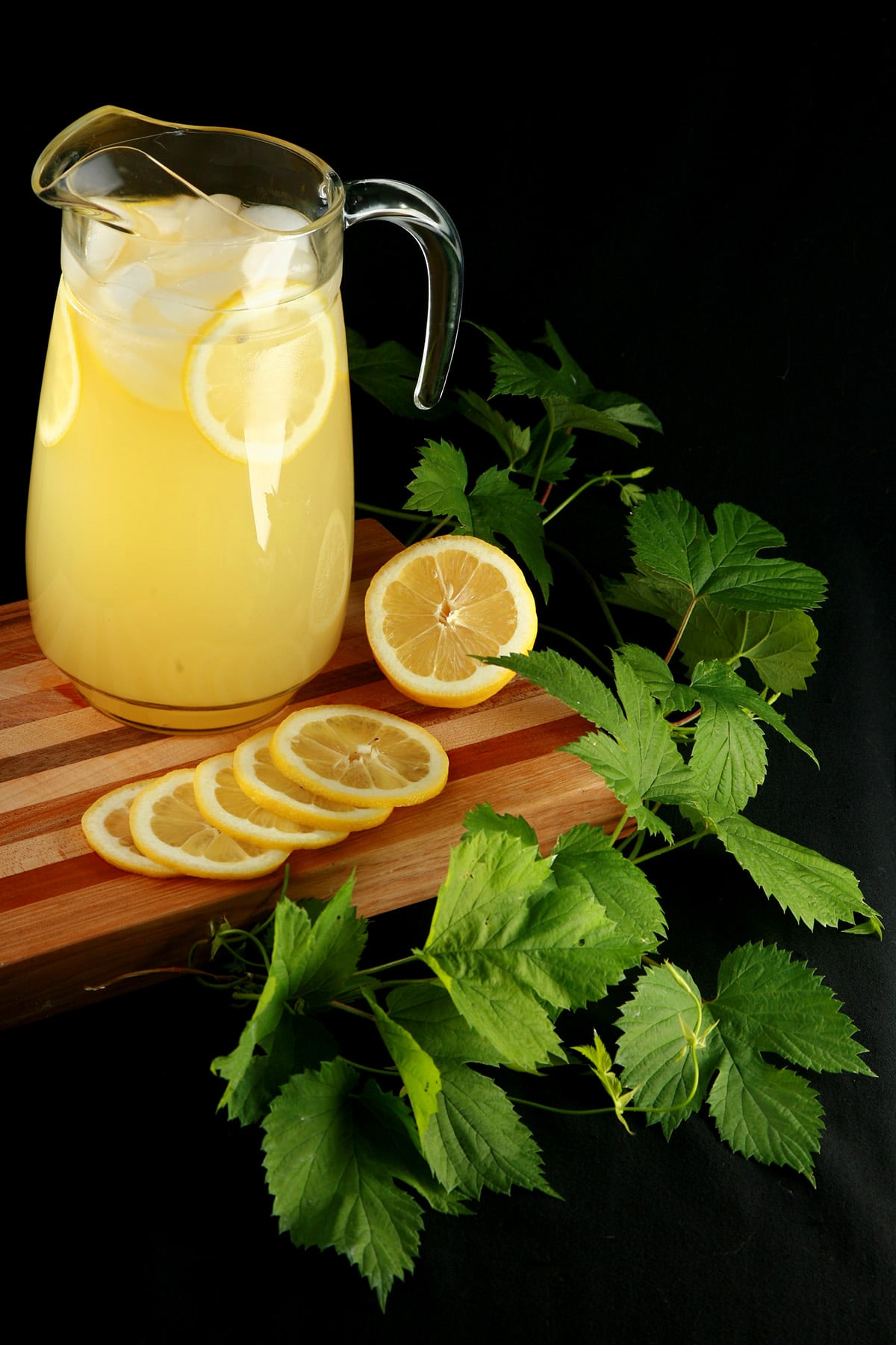 A pitcher of hop lemonade rests on a wooden board, with slices lemons and a hop bine next to it.