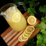 A pitcher of hop lemonade rests on a wooden board, with slices lemons and a hop bine next to it.