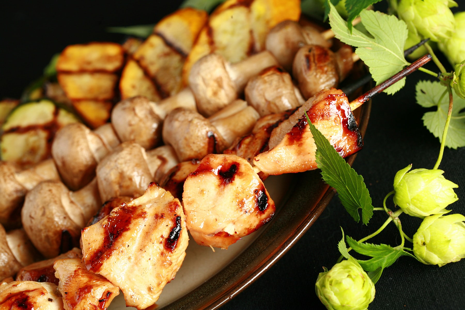 Skewers of hop-marinated grilled chicken, mushrooms, and zucchini are lined up on a brown platter. A hop bine with flowers wraps around the back end of the platter.
