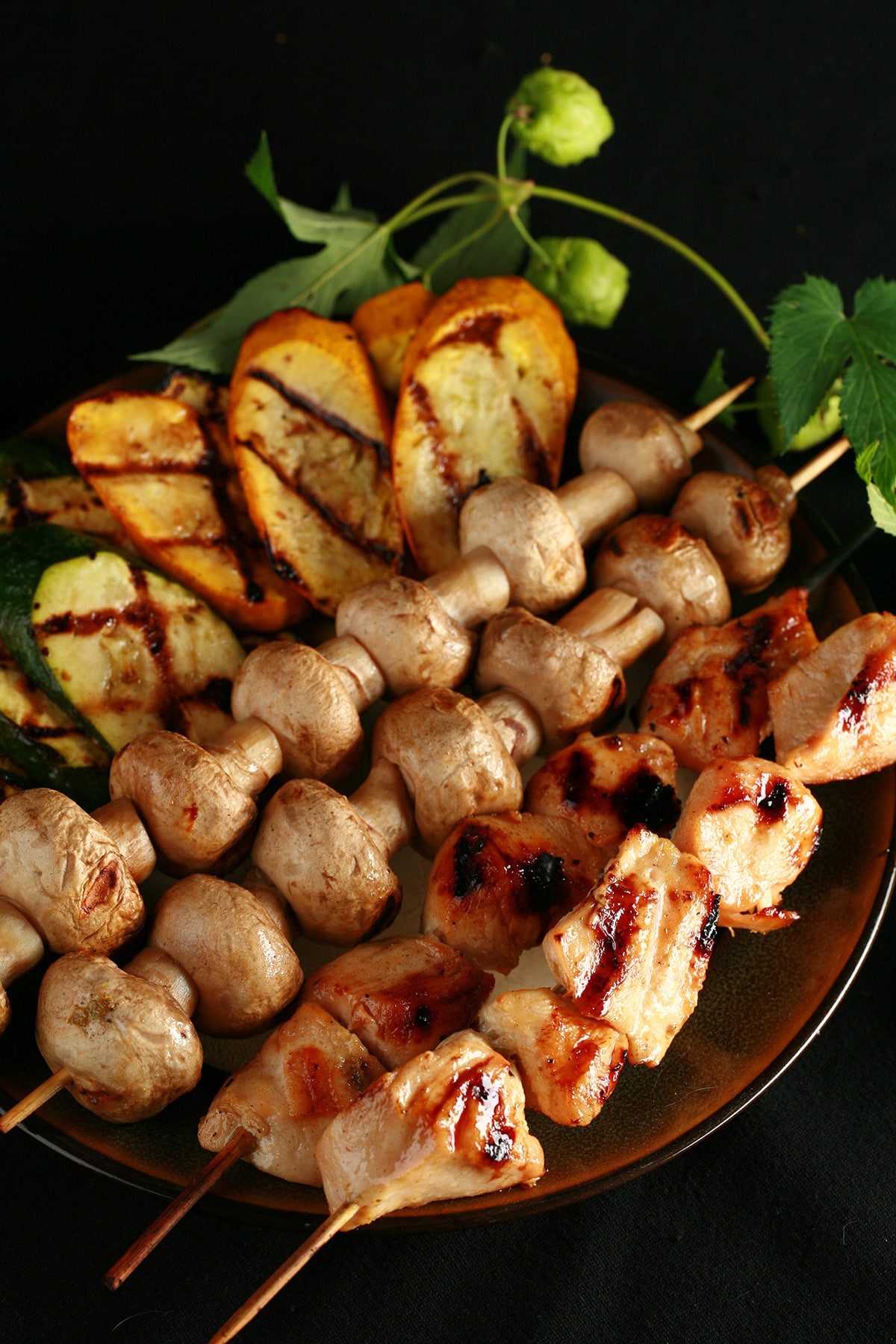 Skewers of hop-marinated grilled chicken, mushrooms, and zucchini are lined up on a brown platter. A hop bine with flowers wraps around the back end of the platter.