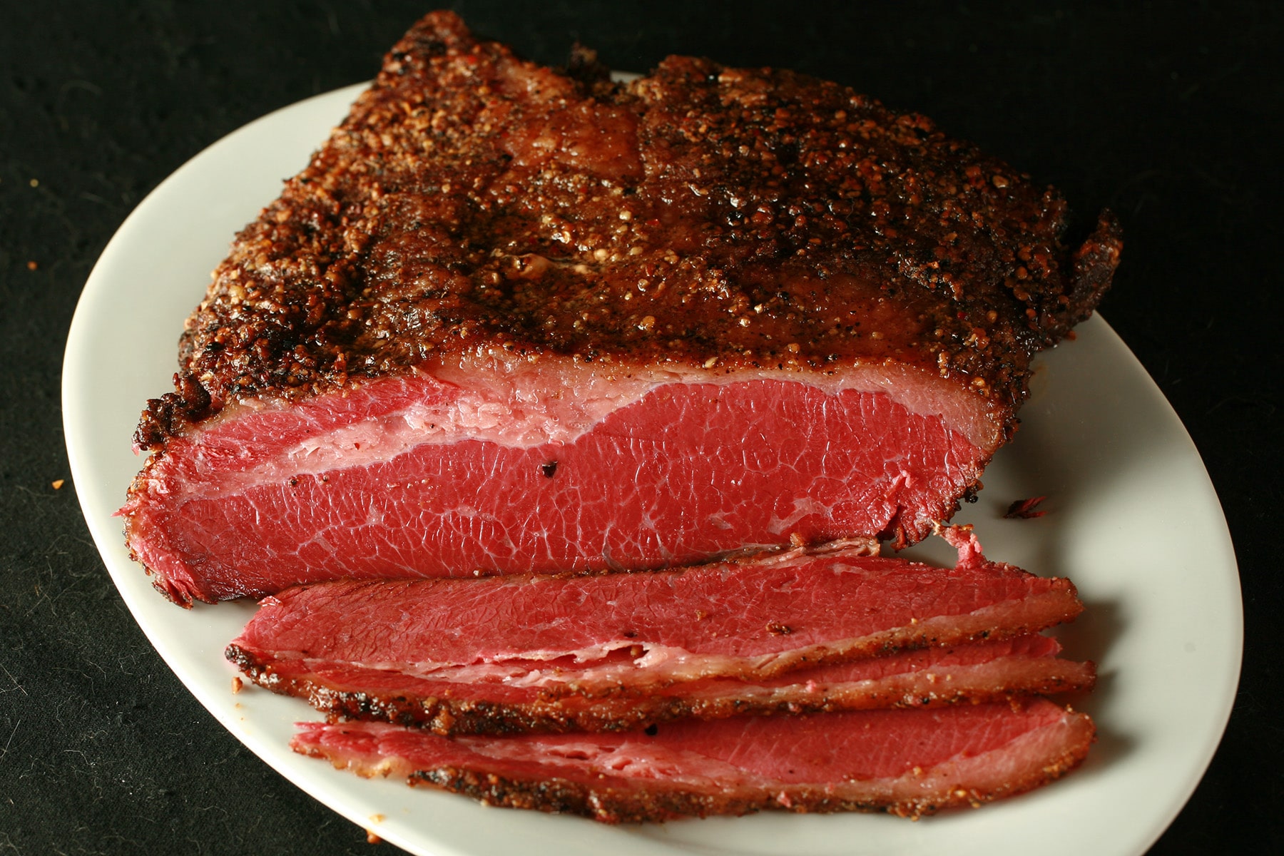 A large slab of Montreal Smoked Meat on a white plate. Several slices have been cut from the end in the foreground, revealing a bright red meat.