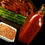 Montreal steak spice in a small square bowl, and marinade in a glass bottle. Behind them, a steak with sauteed mushrooms and asparagus on a plate.