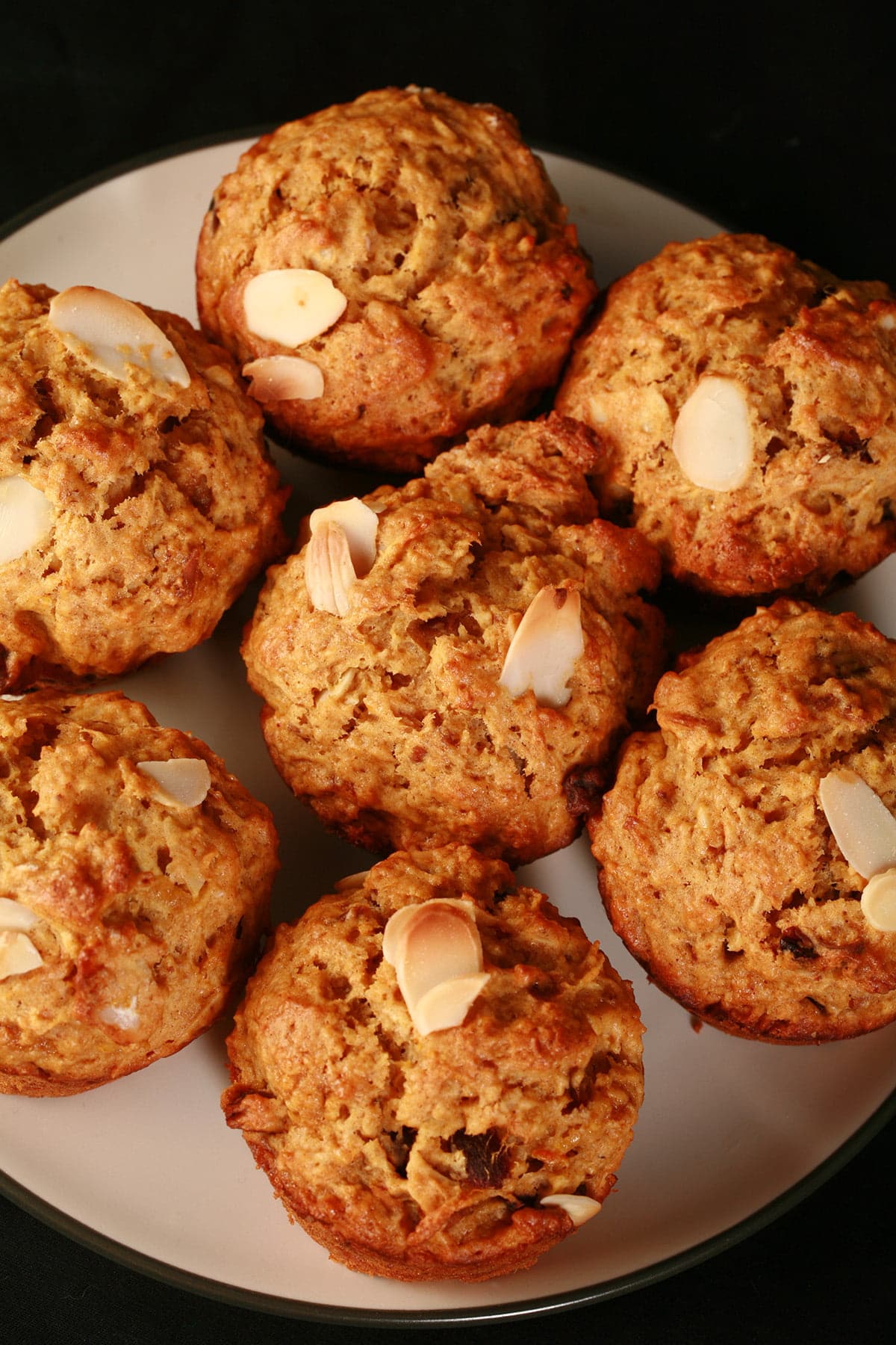 A plate of orange date and almond muffins. They are an orangey brown colour, with slices of almonds and chunks of dates visible throughout.