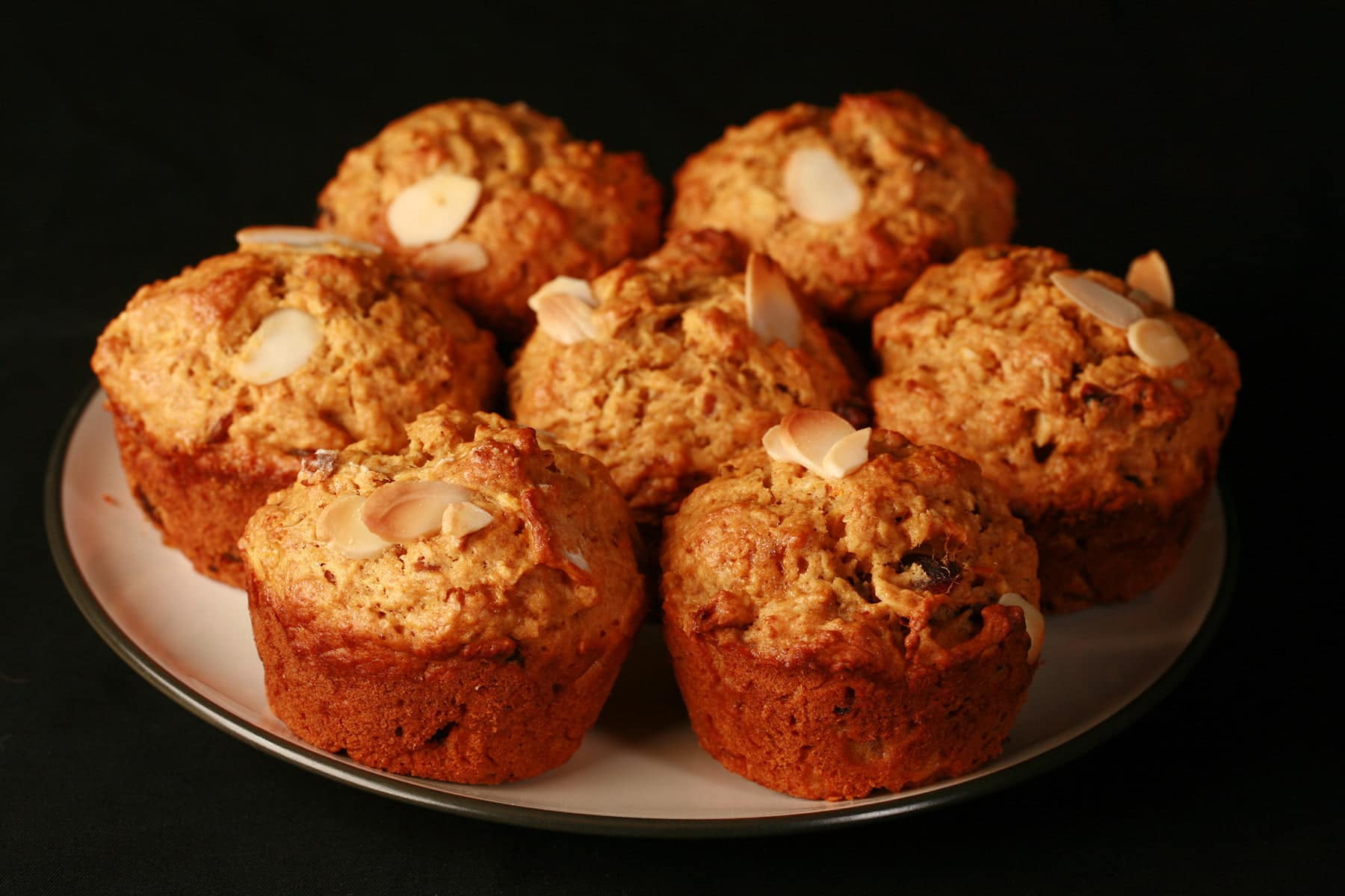 A plate of orange-brown muffins, with slices of almonds and chunks of dates visible throughout.