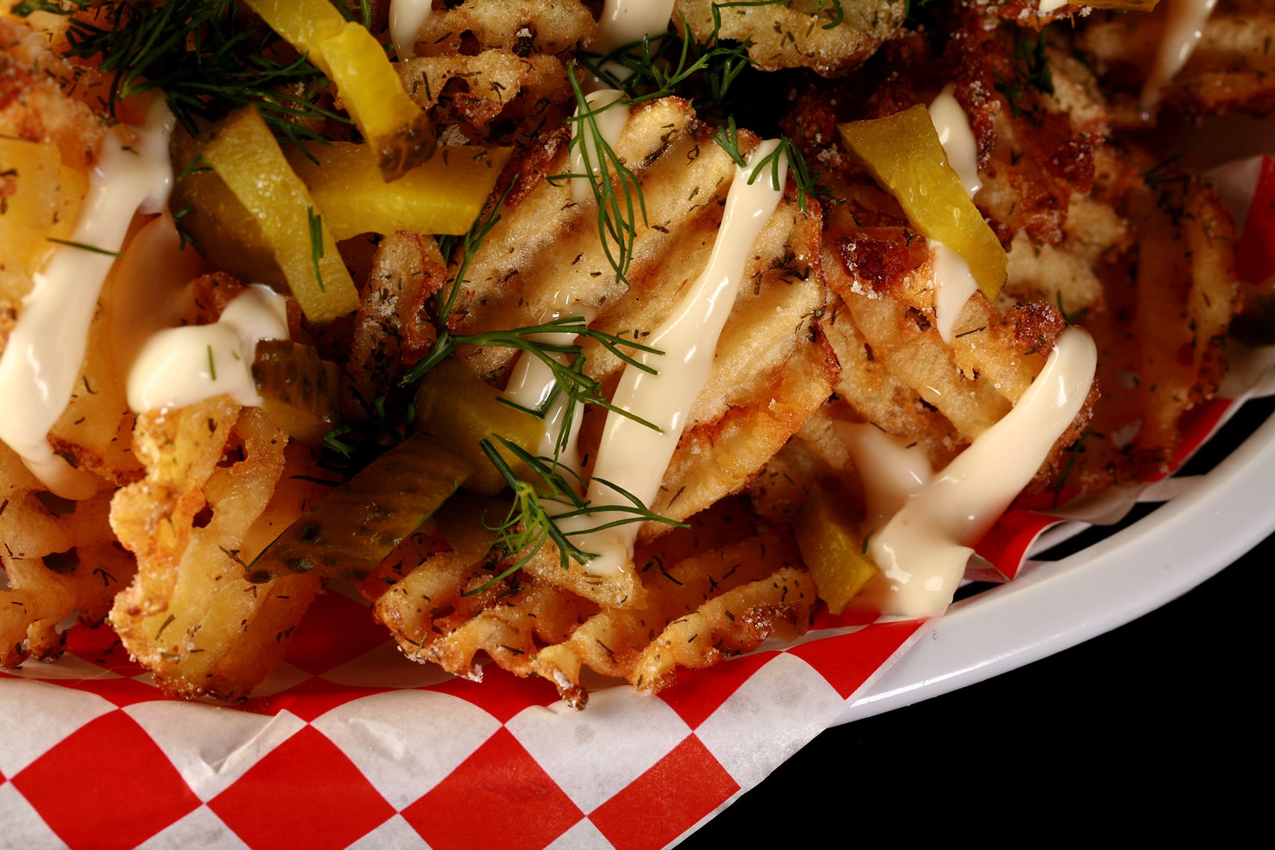 Waffle cut fries, in a white basket lined with red and white checkered paper. The fries are drizzled with roasted garlic aioi and topped with chopped dill and chopped pickles.