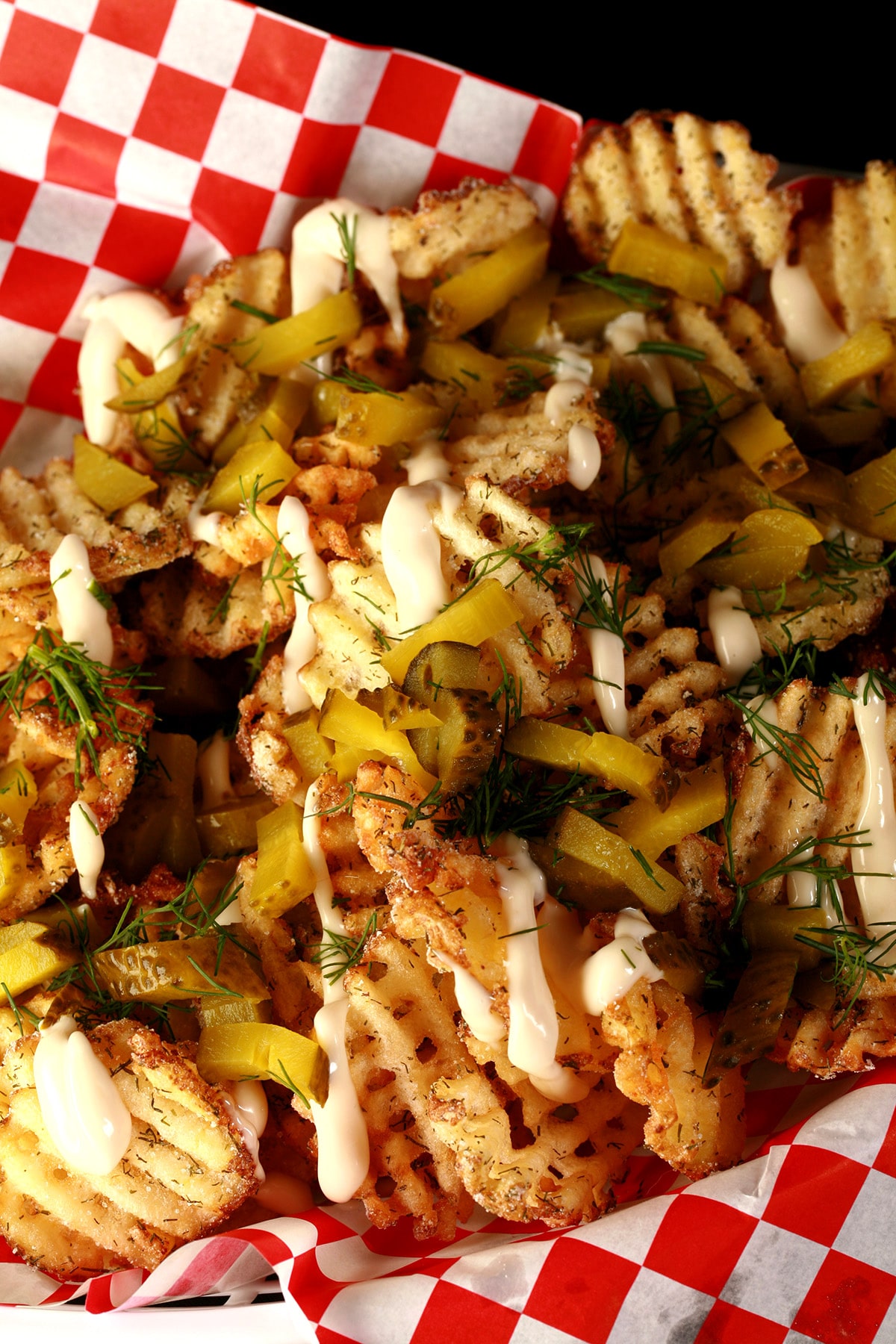 Dill Pickle waffle fries, in a white basket lined with red and white checkered paper. The fries are drizzled with roasted garlic aioi and topped with chopped dill and chopped pickles.
