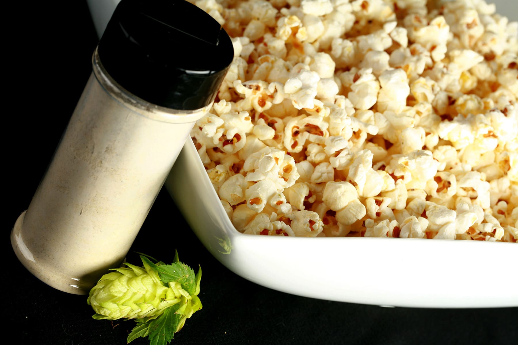 A large rectangular bowl holds hopped popcorn - hopcorn! There is a small canister of the popcorn seasoning, and a fresh hop flower next to the bowl.