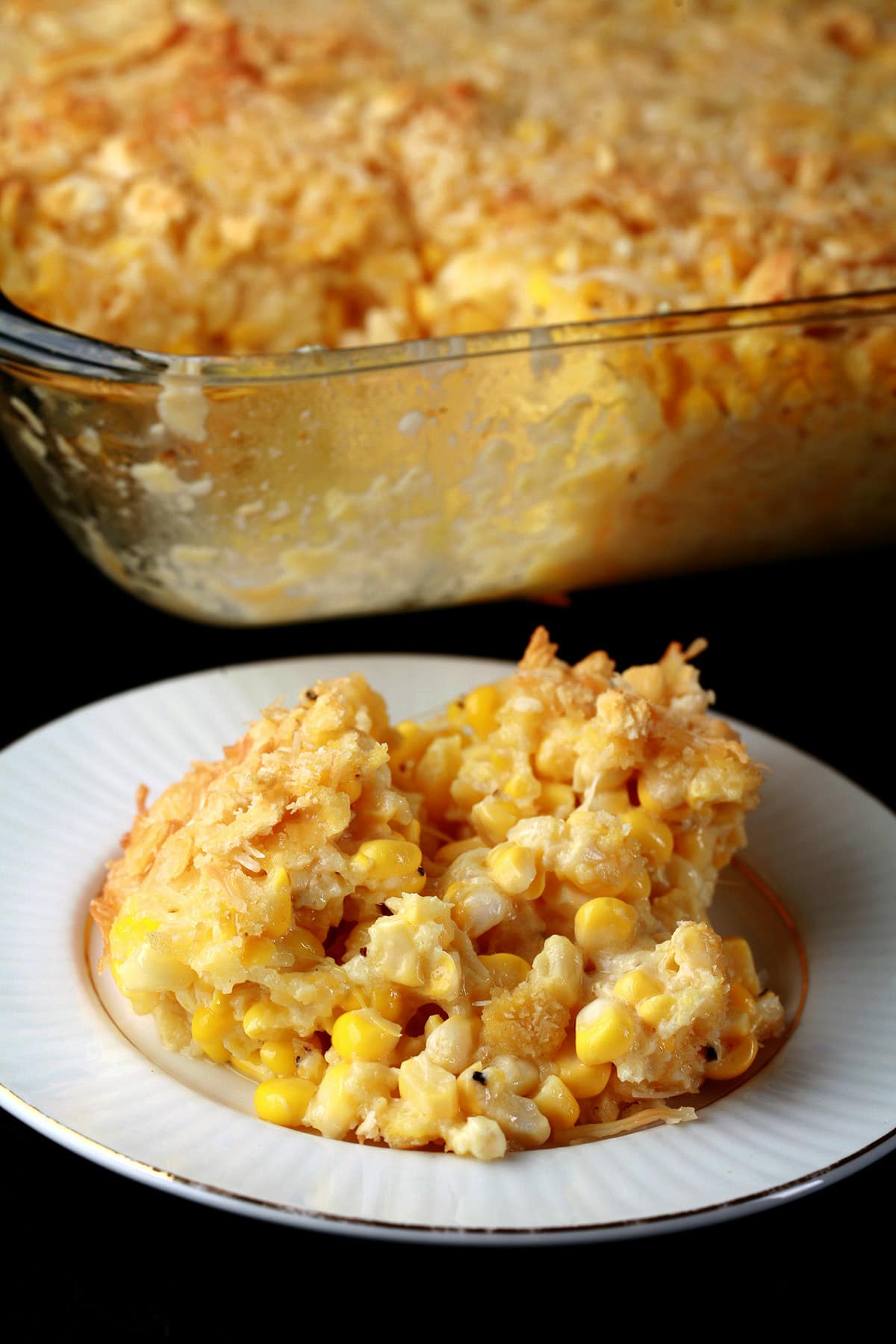 A serving of scalloped corn - fresh sweet corn, cheese, crackers, and more - on a small white plate. The pan of casserole it was served from sits behind the plate.