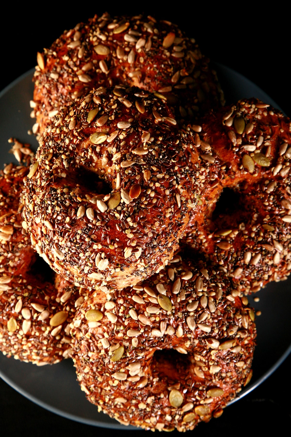 A plate stacked with a pile of whole wheat and flax bagels. The top side of each bagel is covered in a mix of seeds - pumpkin, sunflower, flax, sesame, chia, and poppy.