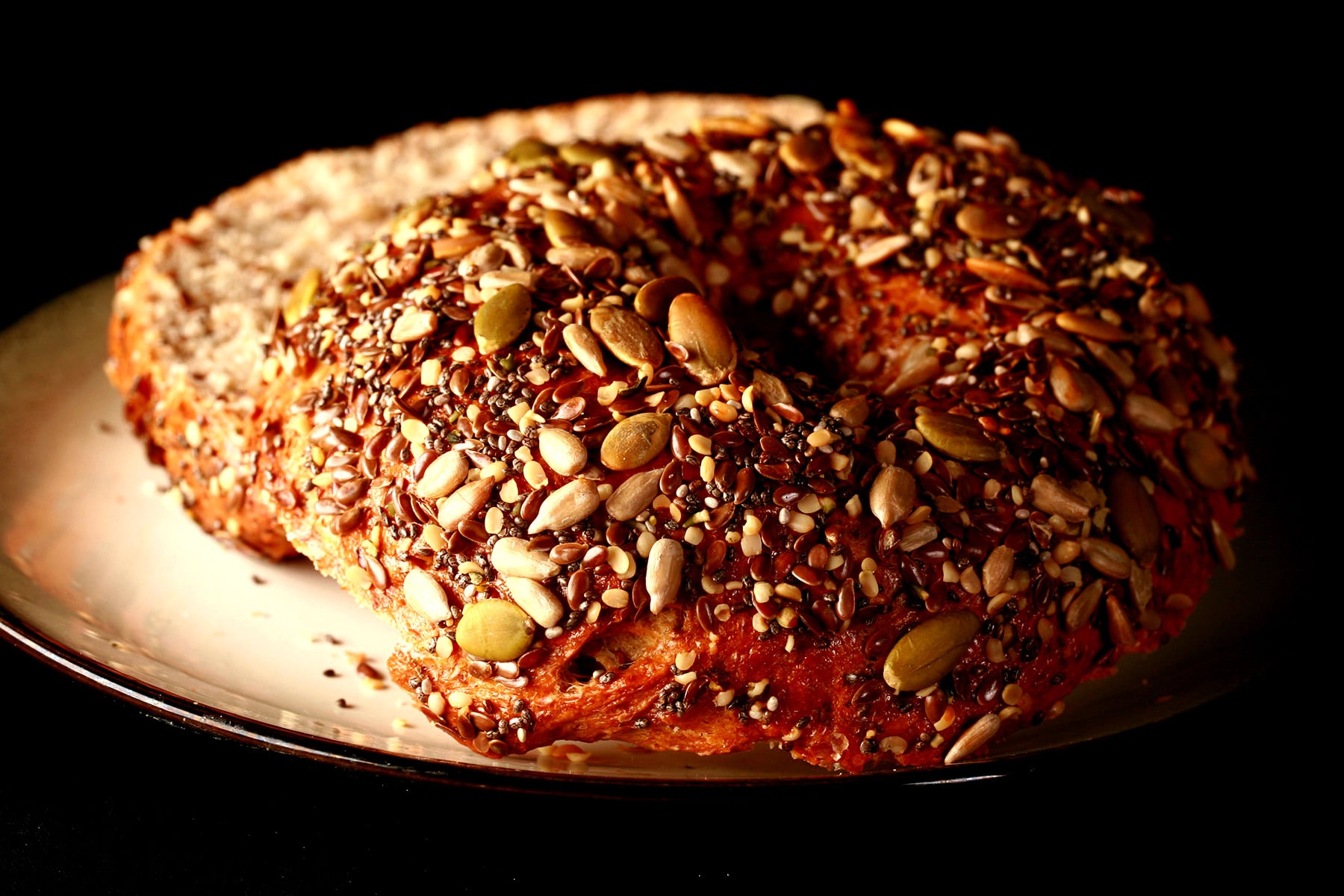 A whole wheat and flax bagel, sliced and served on a small plate. The top side of the bagel is covered in a mix of seeds - pumpkin, sunflower, sesame, chia, and poppy.