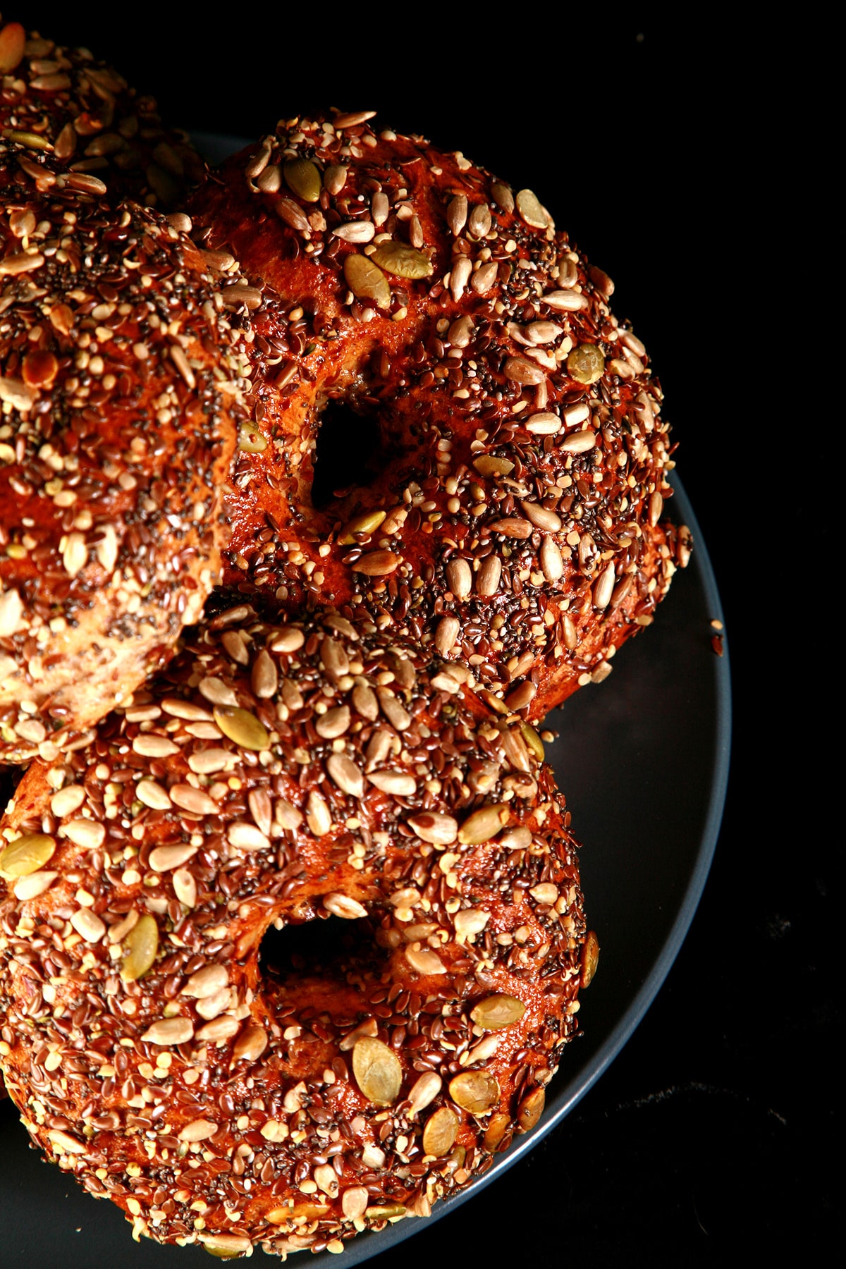 A plate stacked with a pile of whole wheat and flax bagels. The top side of each bagel is covered in a mix of seeds - pumpkin, sunflower, flax, sesame, chia, and poppy.