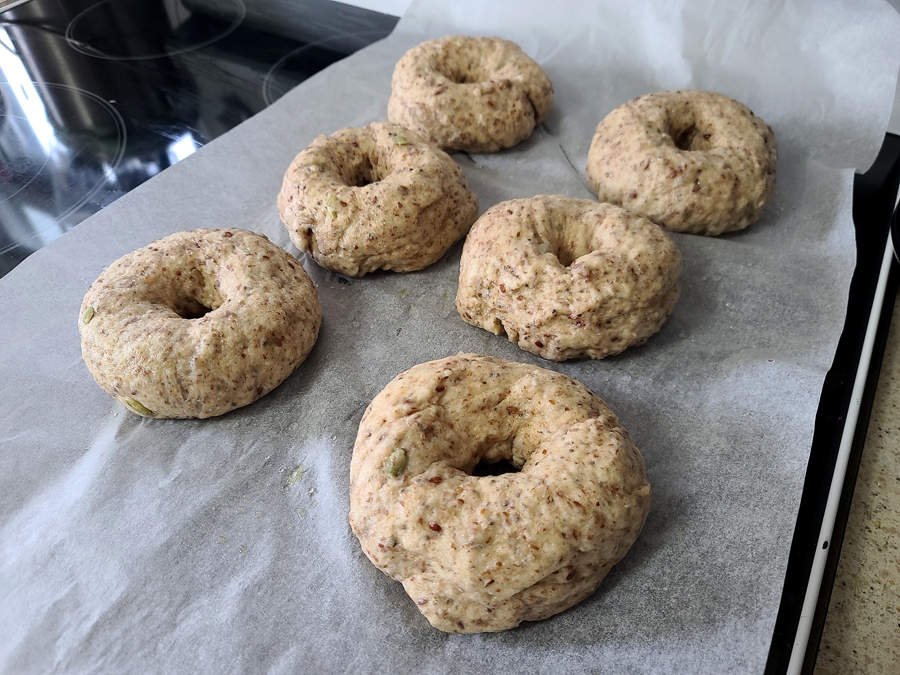 6 boiled seeded whole wheat flax bagels resting on a parchment lined baking sheet.