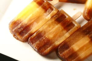 Several frozen root beer popsicles with a stripe of orange soda in the middle - are laid out on a white plate. These are a homemade version of "Space Shuttle" popsicles, a discontinued Dickie Dee item.