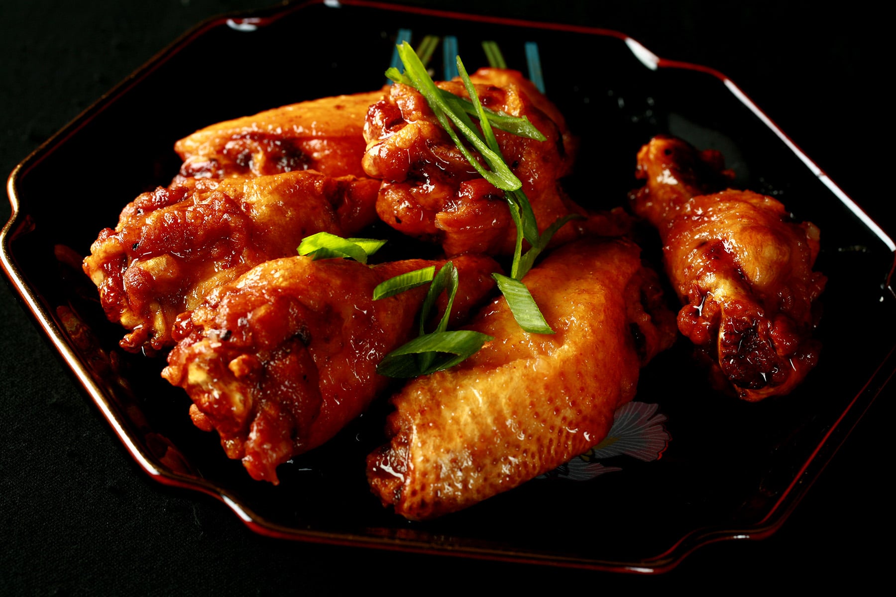 A small black plate with a pile of Spicy Ginger Wings piled on it, topped with slices of green onion.